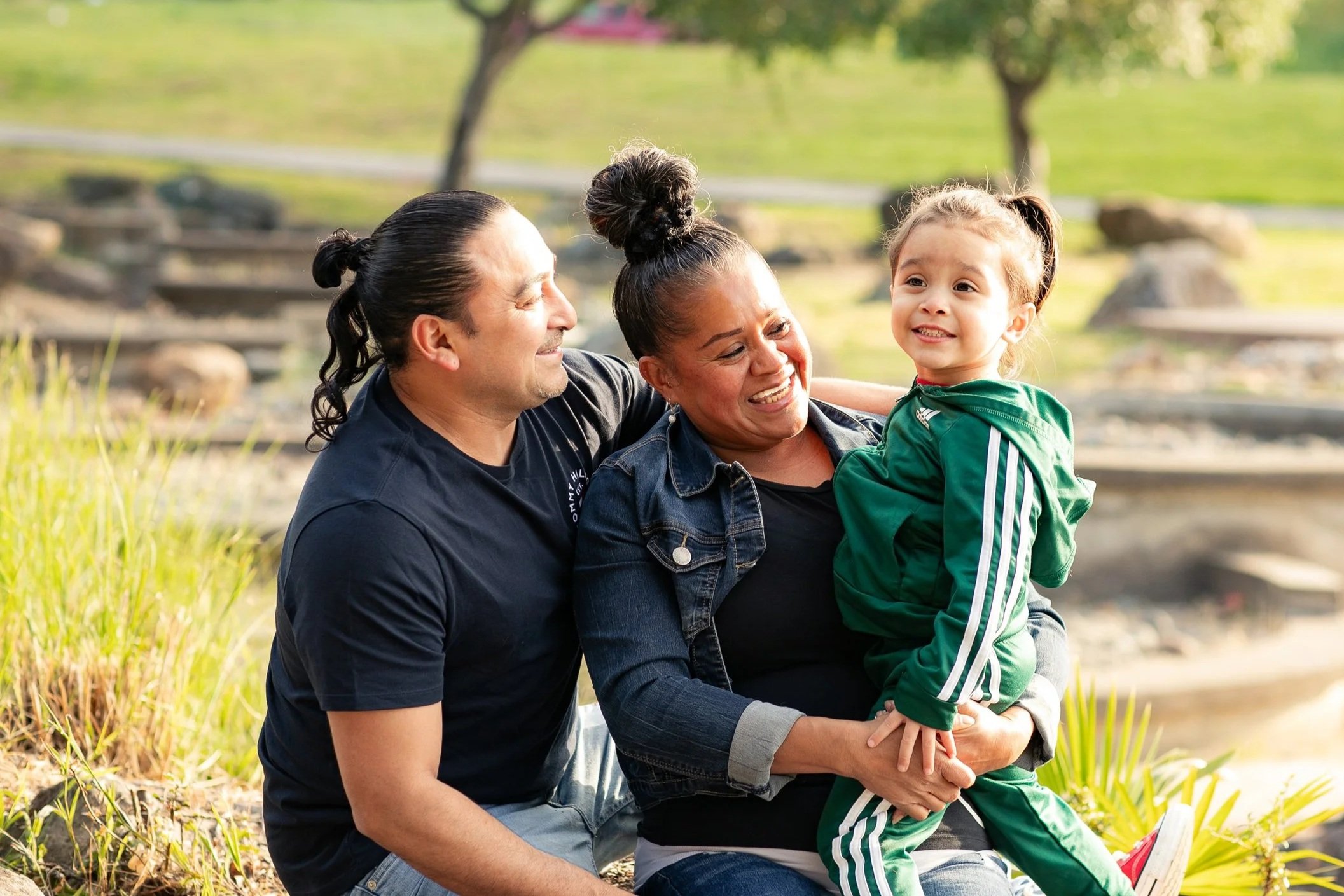 A happy family of three, including a father, mother, and young daughter, enjoying outdoors in a park during daytime.