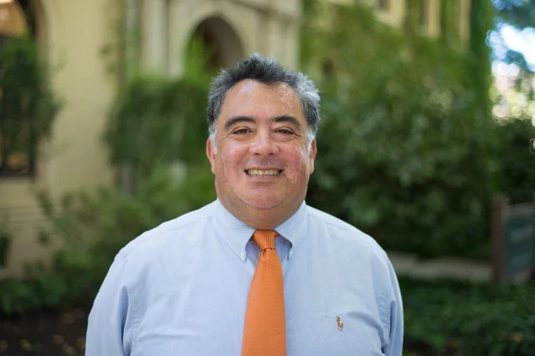 A smiling man in a light blue dress shirt and orange tie standing outdoors with green trees and a building in the background.