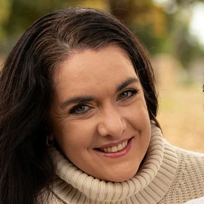 Close-up of a woman smiling outdoors with trees in the background, wearing a beige turtleneck sweater.