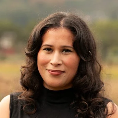 A woman with dark, curly hair wearing a sleeveless black top outdoors with a blurred natural background.