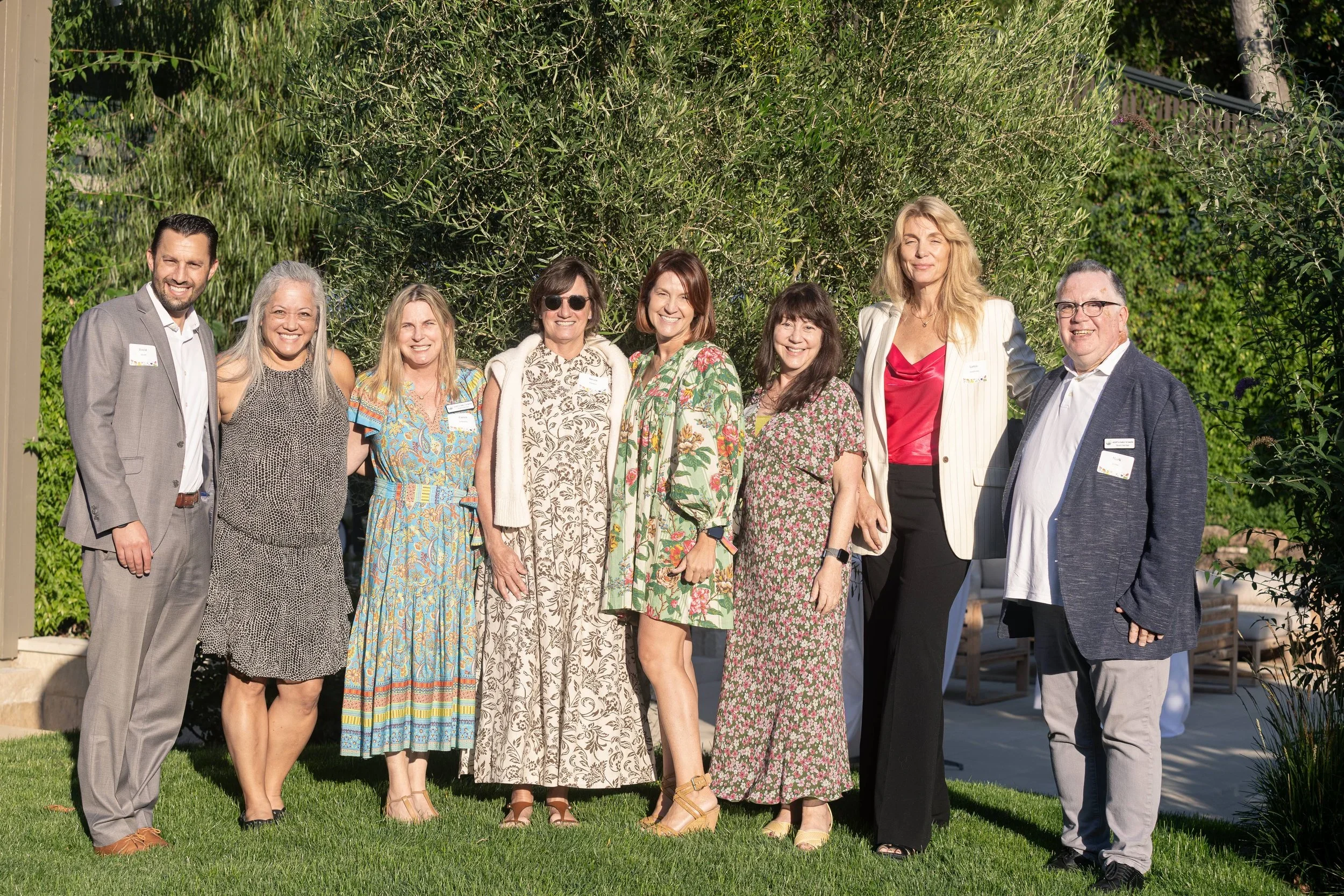 Group of nine diverse adults smiling and standing outdoors in front of trees and a garden, dressed in casual to business casual attire.