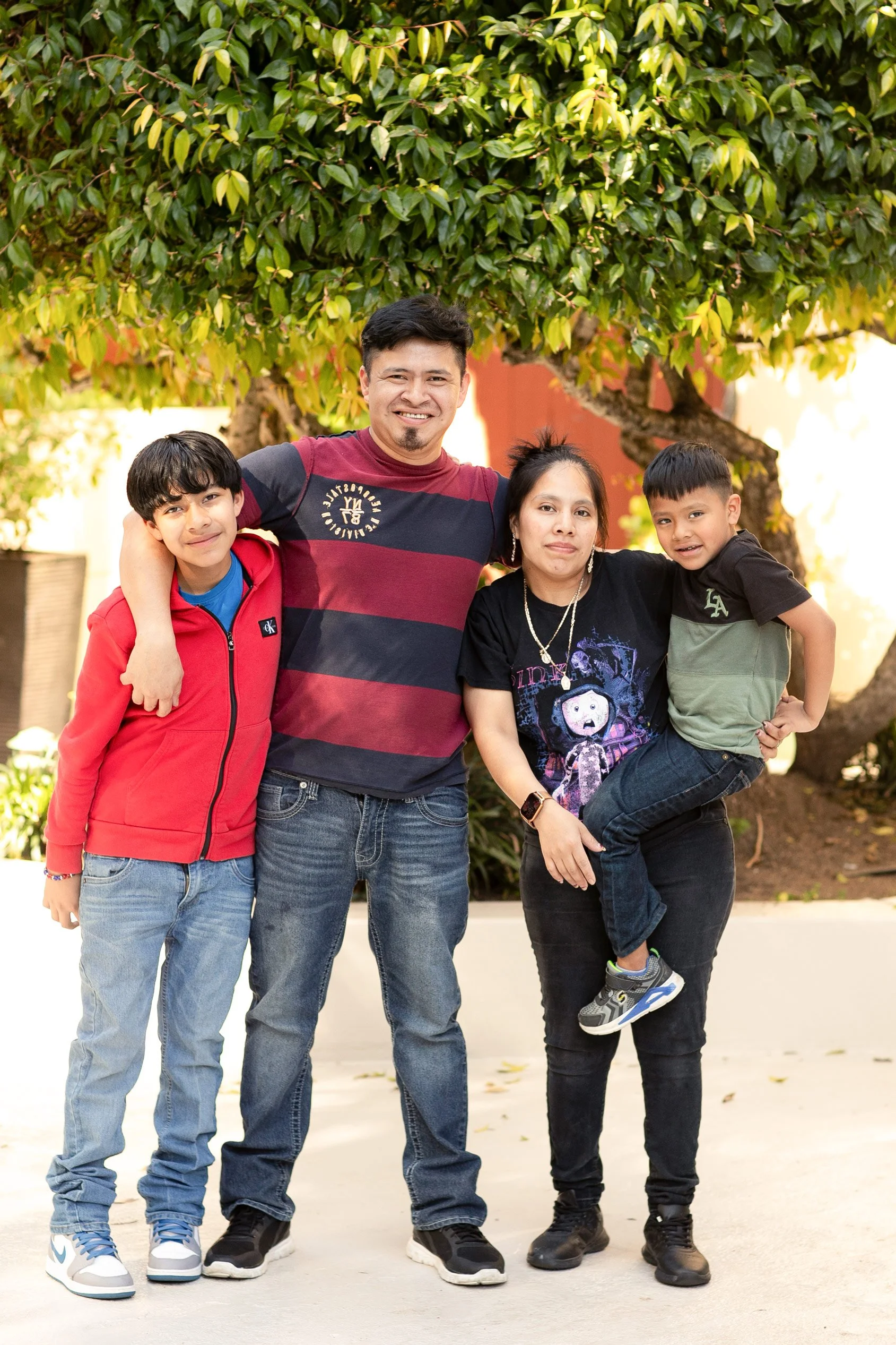 A family of five smiling and standing outdoors in front of a tree. The group includes two adult women, an adult man, and two young boys. They are dressed casually and appear happy.