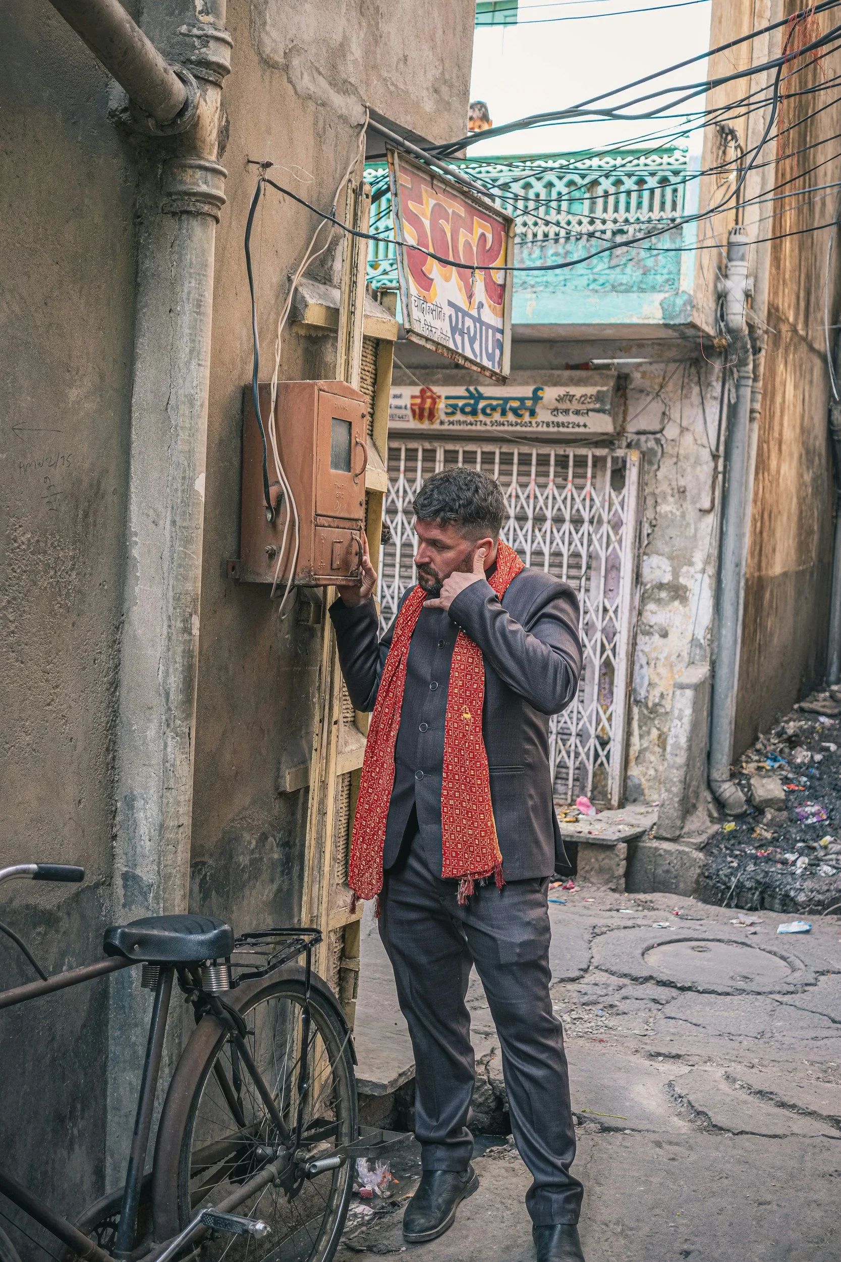 A man in a gray suit with a red scarf talking on a mobile phone in an alleyway. There is a black bicycle with a basket parked against the wall, and electrical wires hanging overhead. The alley has a metal gate and some scattered trash. The man appears to be focused on his call.