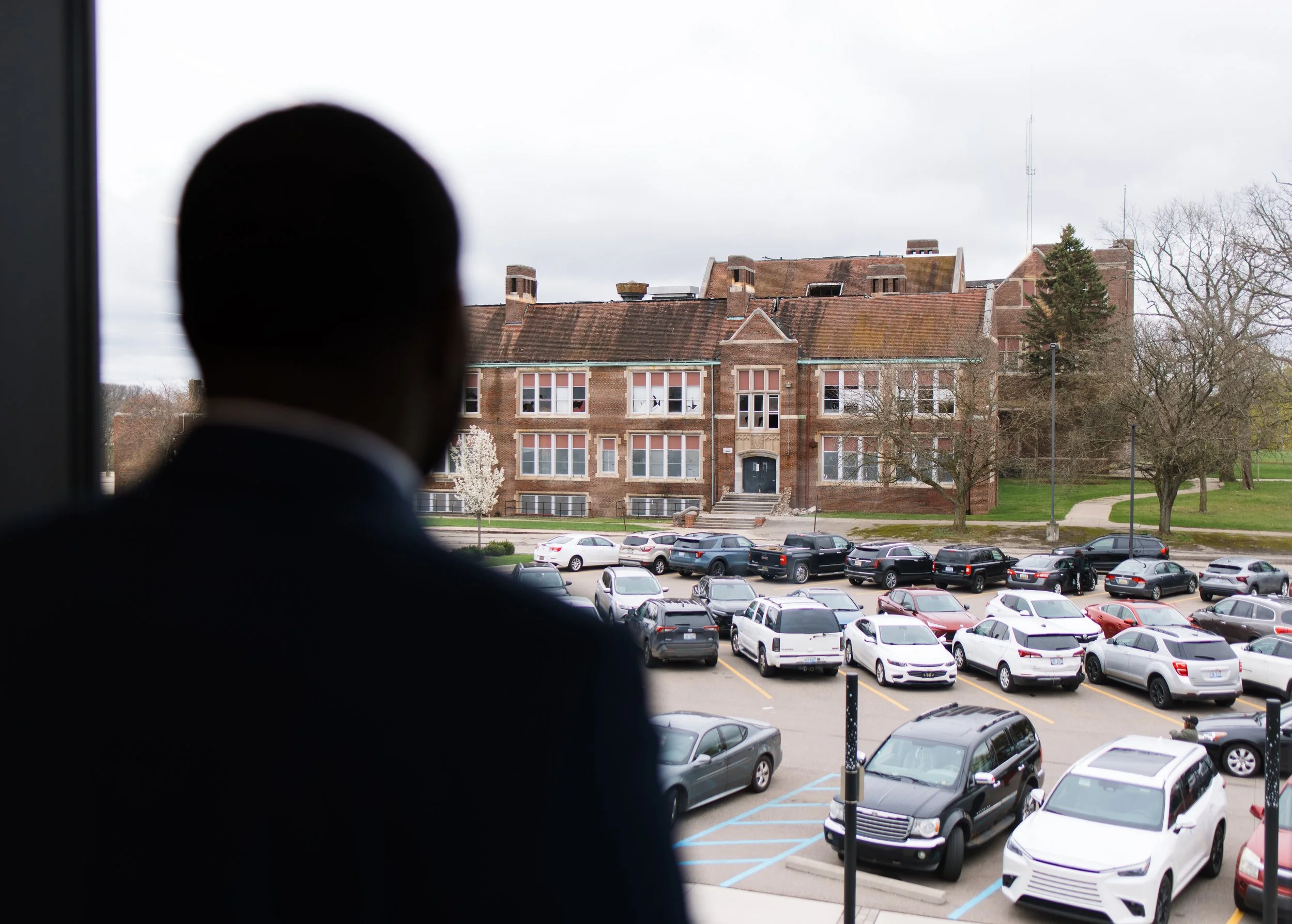 A person looking out of a window at a parking lot with multiple cars and a large brick building with a sloped roof, trees, and a cloudy sky in the background.