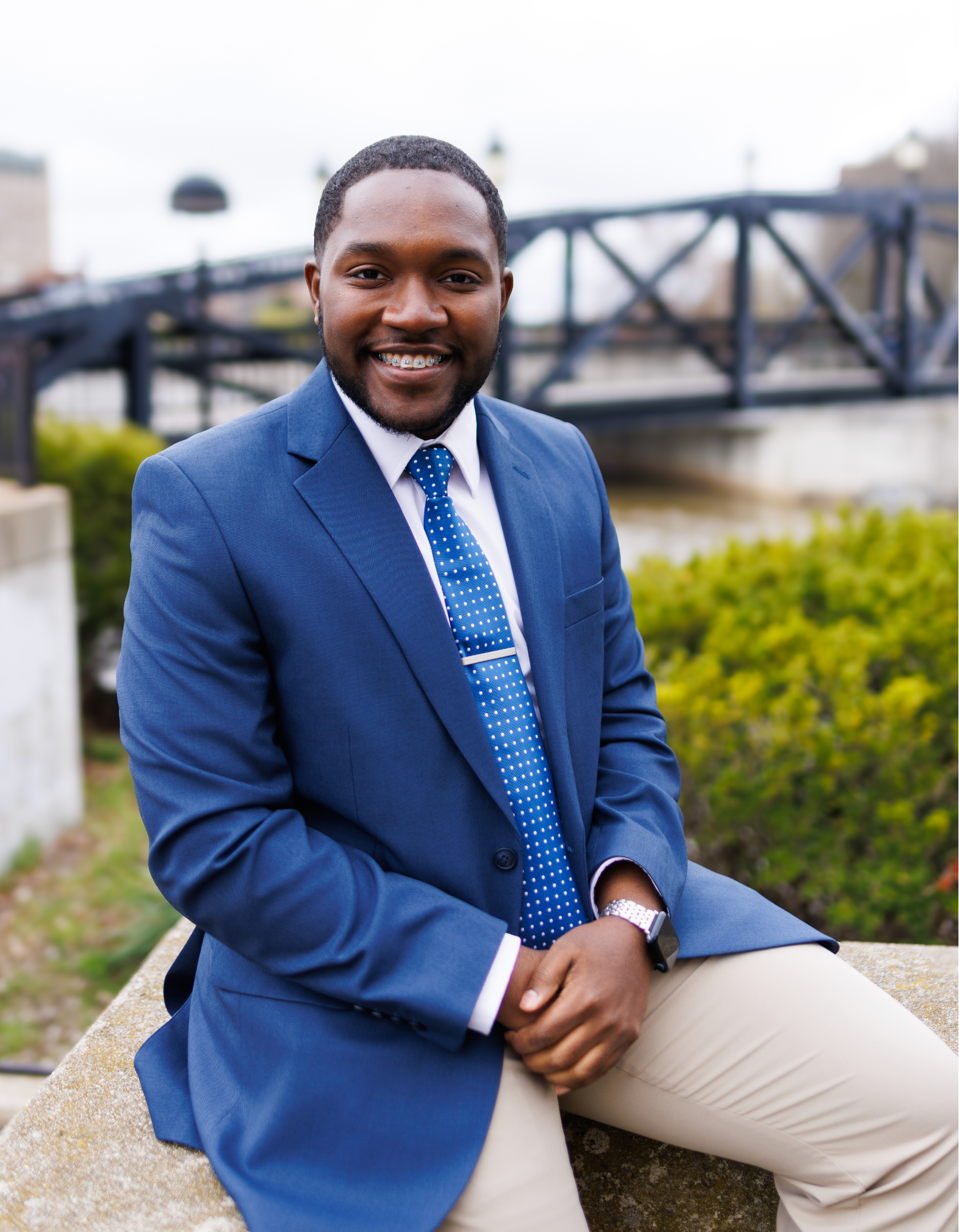 A young man in a navy blue blazer, white shirt, and blue polka dot tie is sitting outdoors on a stone surface. He has a friendly smile with braces and is wearing a watch. The background shows a bridge over water and greenery.