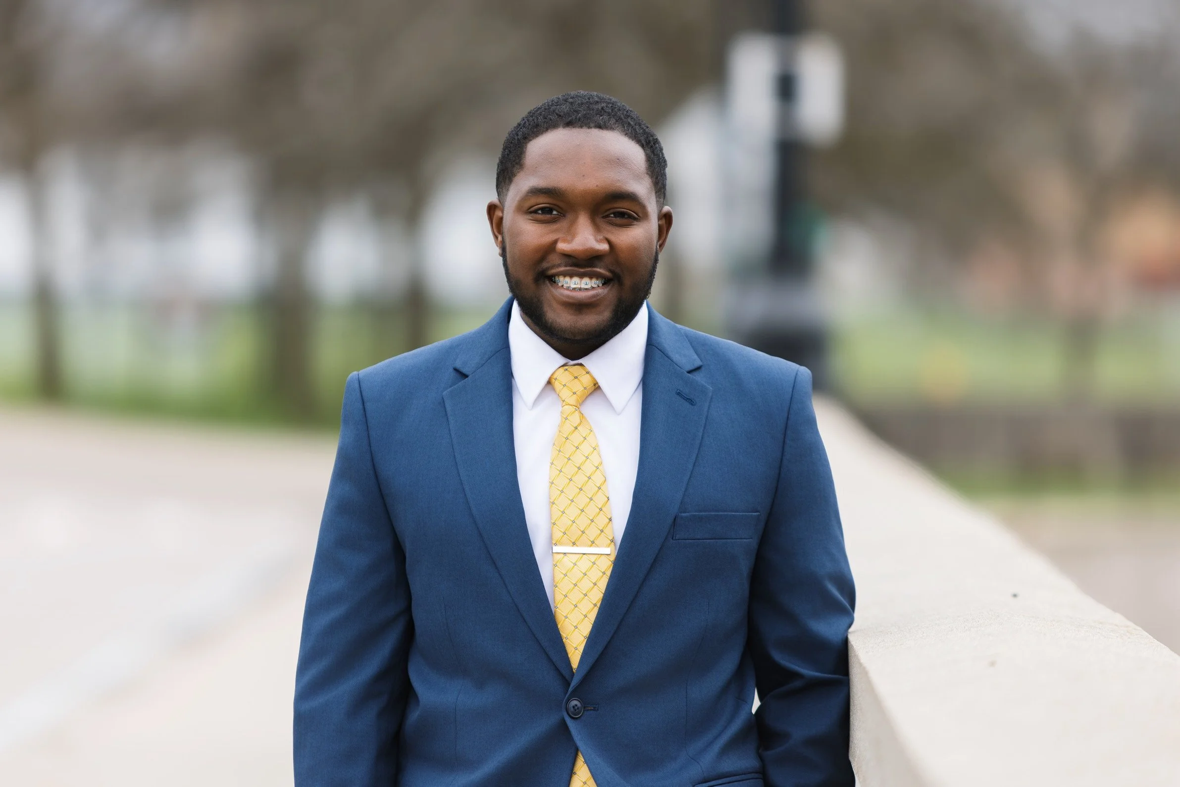 A young man wearing a blue suit, white shirt, and yellow tie, smiling with braces, standing outdoors near a stone railing, with blurred trees and park background.