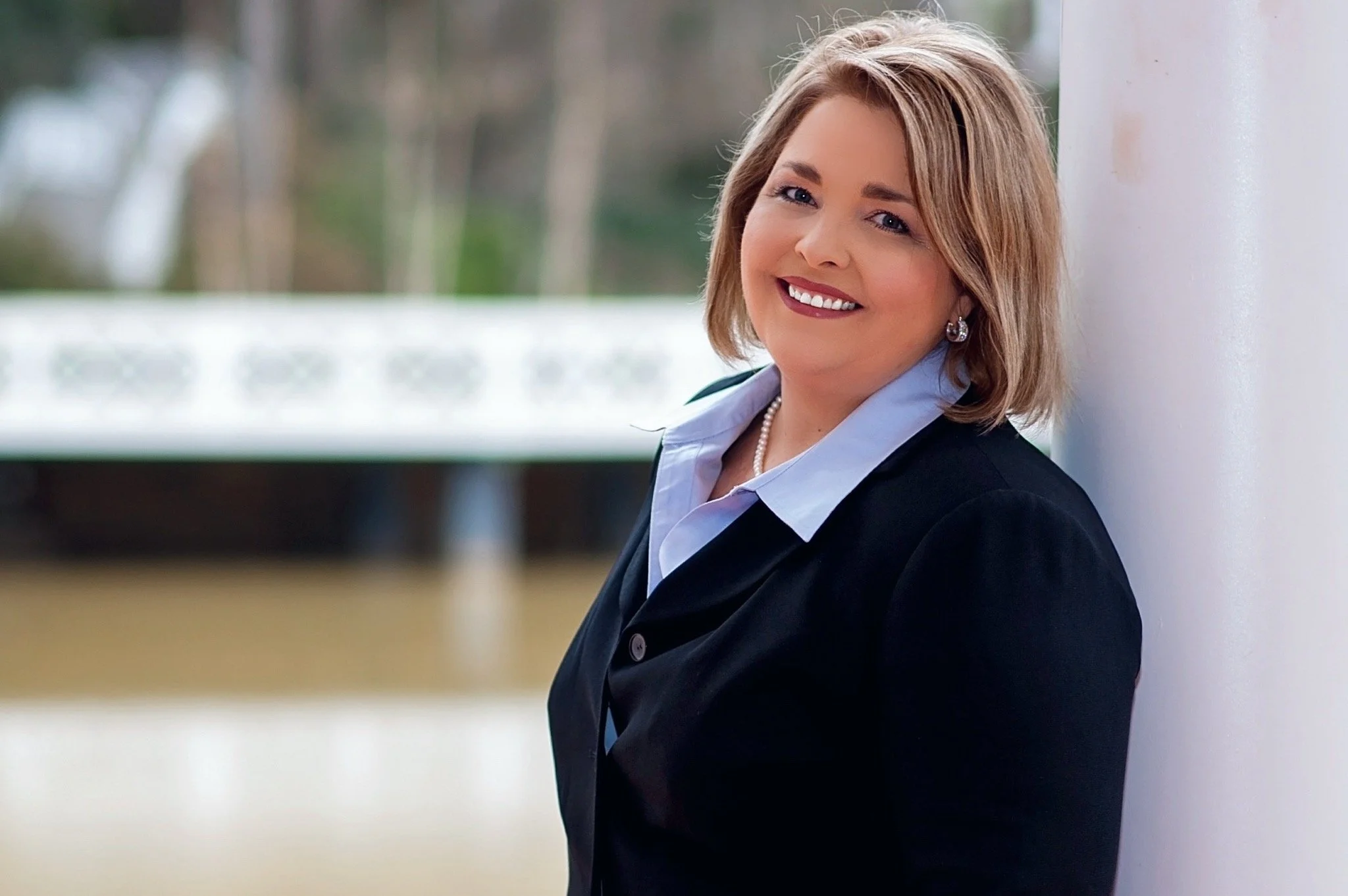 A woman with blonde hair smiling, leaning against a white column indoors, wearing a dark blazer, light blue shirt, pearl necklace, and earrings.