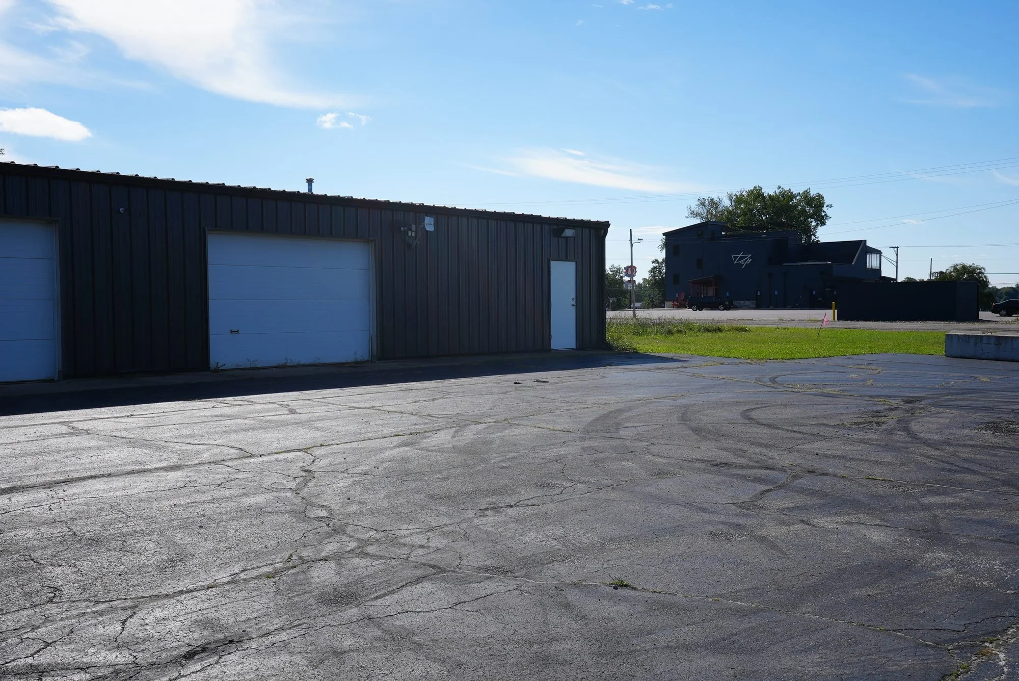 A large paved parking lot with visible cracks in the surface, next to a black metal building with white garage doors and a side door. In the background, there is a modern black building with some trees and power lines against a partly cloudy sky.