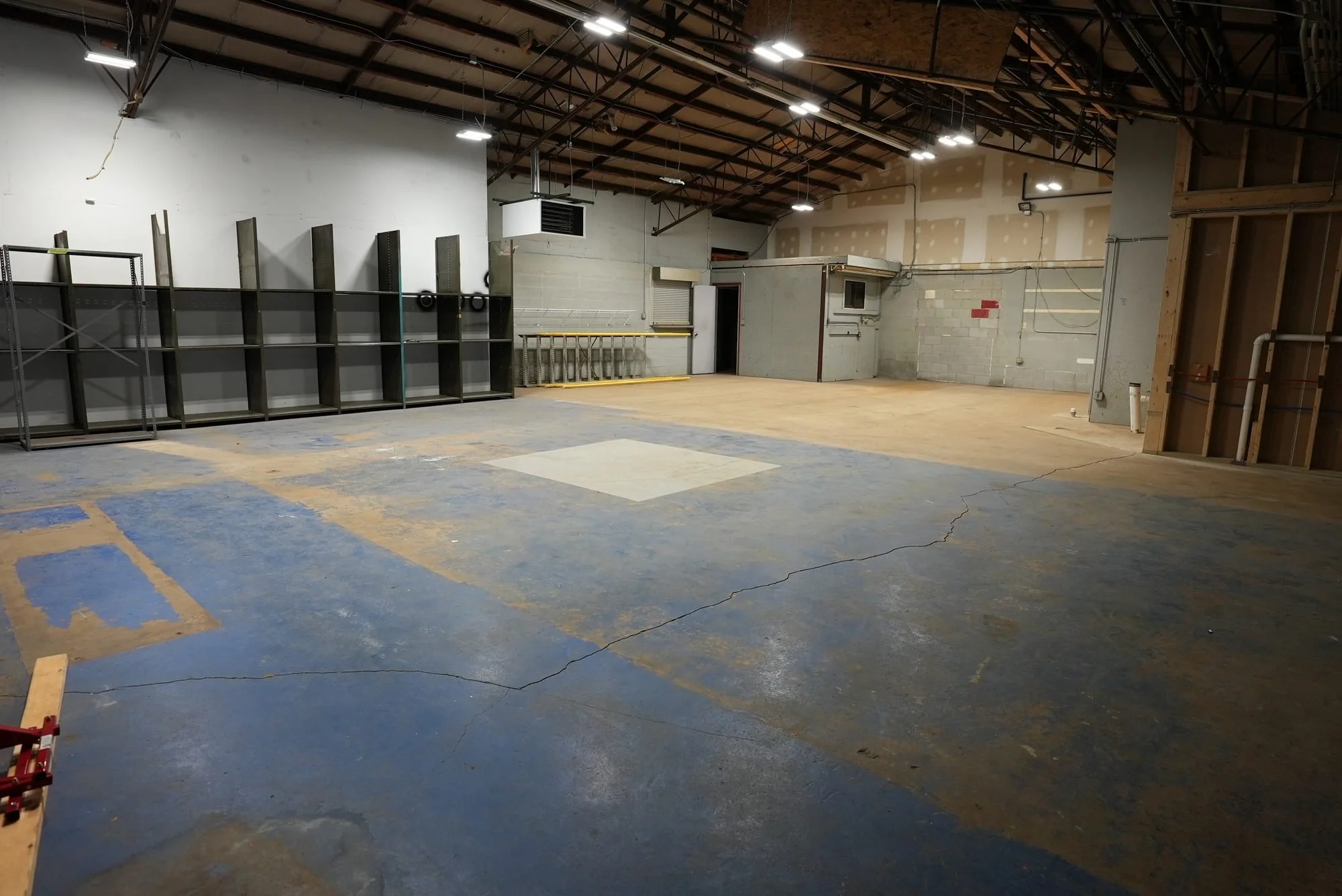 Empty indoor warehouse with a cracked concrete floor, metal shelving units on the left, unfinished walls, exposed ceiling beams, and scattered construction materials.