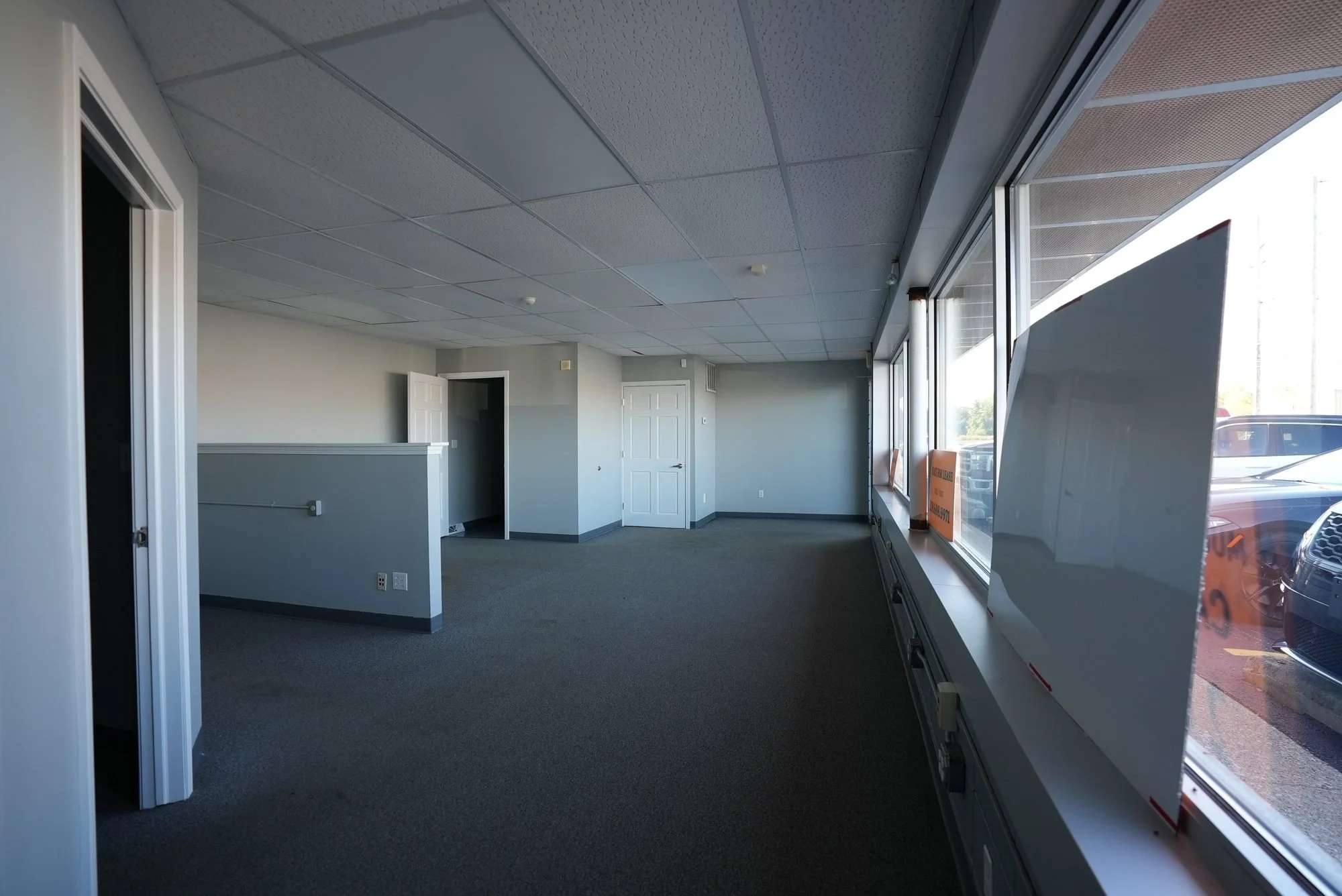 Interior of an empty commercial space with large windows, white walls, tiled ceiling, and gray carpet, with a white door and a sign visible outside.
