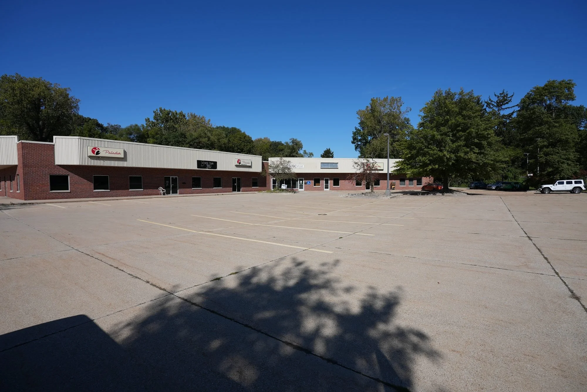 Empty parking lot in front of a single-story commercial building with a sign that reads 'Paladin,' surrounded by trees and a clear blue sky.