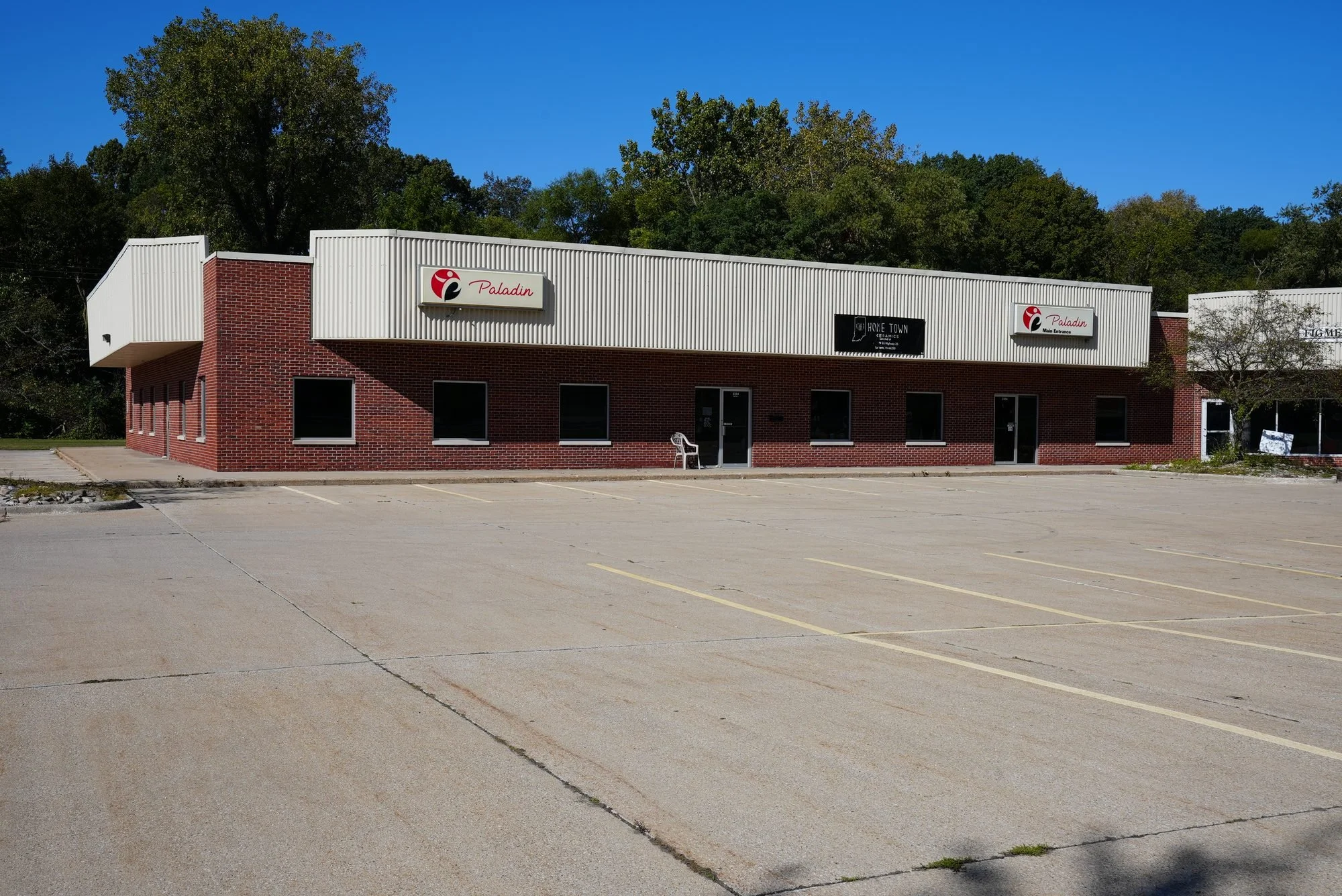 An empty parking lot in front of a commercial building with signs for Paladin and Home Town Ceramics, surrounded by trees under a clear blue sky.