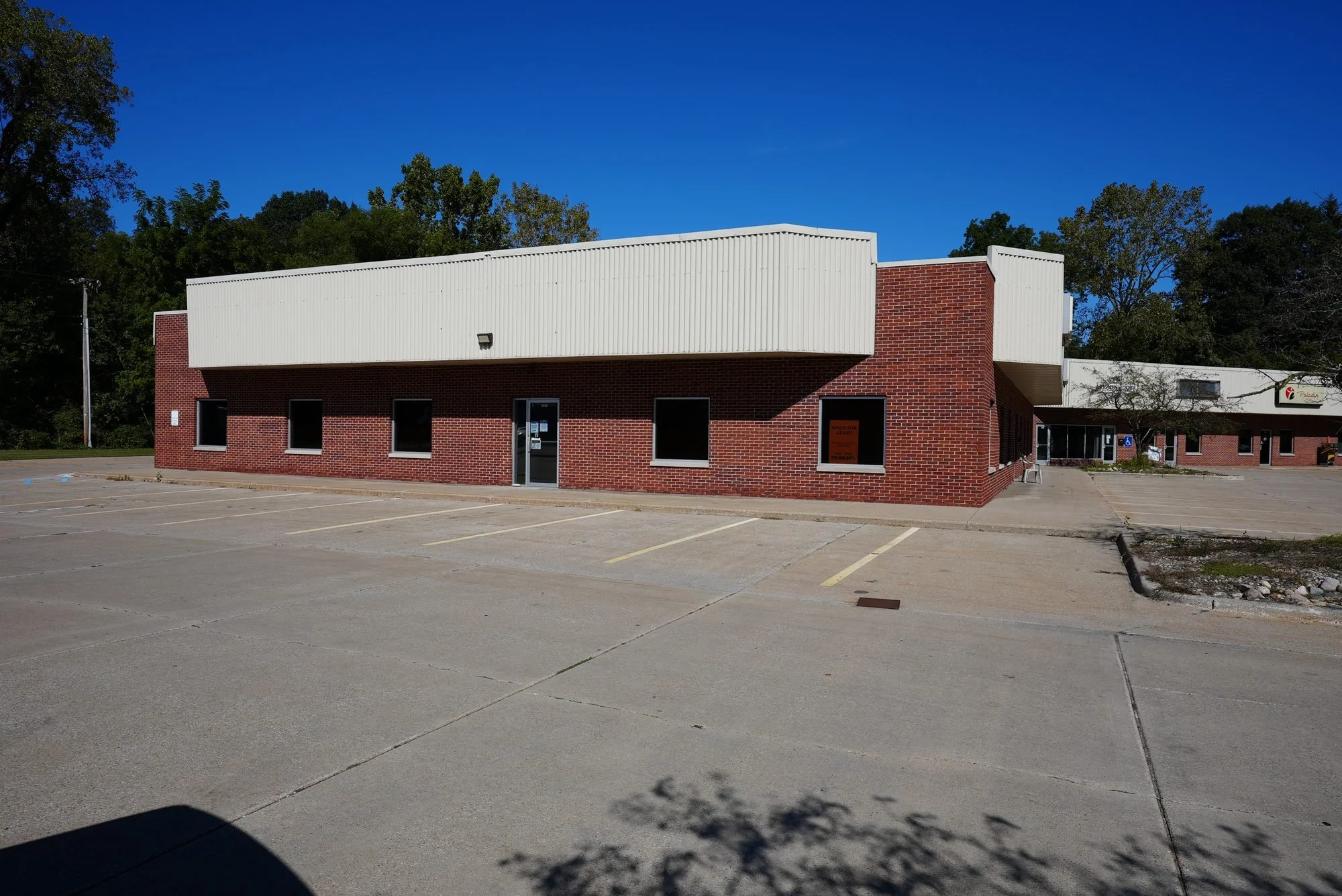 Empty parking lot in front of a brick building with a white overhang, trees in the background, and a clear blue sky.