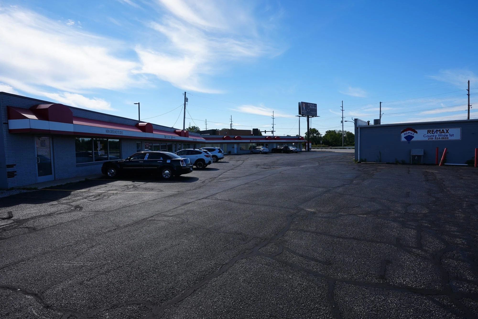 A parking lot outside a commercial shopping plaza with a row of storefronts that have red and white awnings, a few parked cars, and a sky with light clouds.
