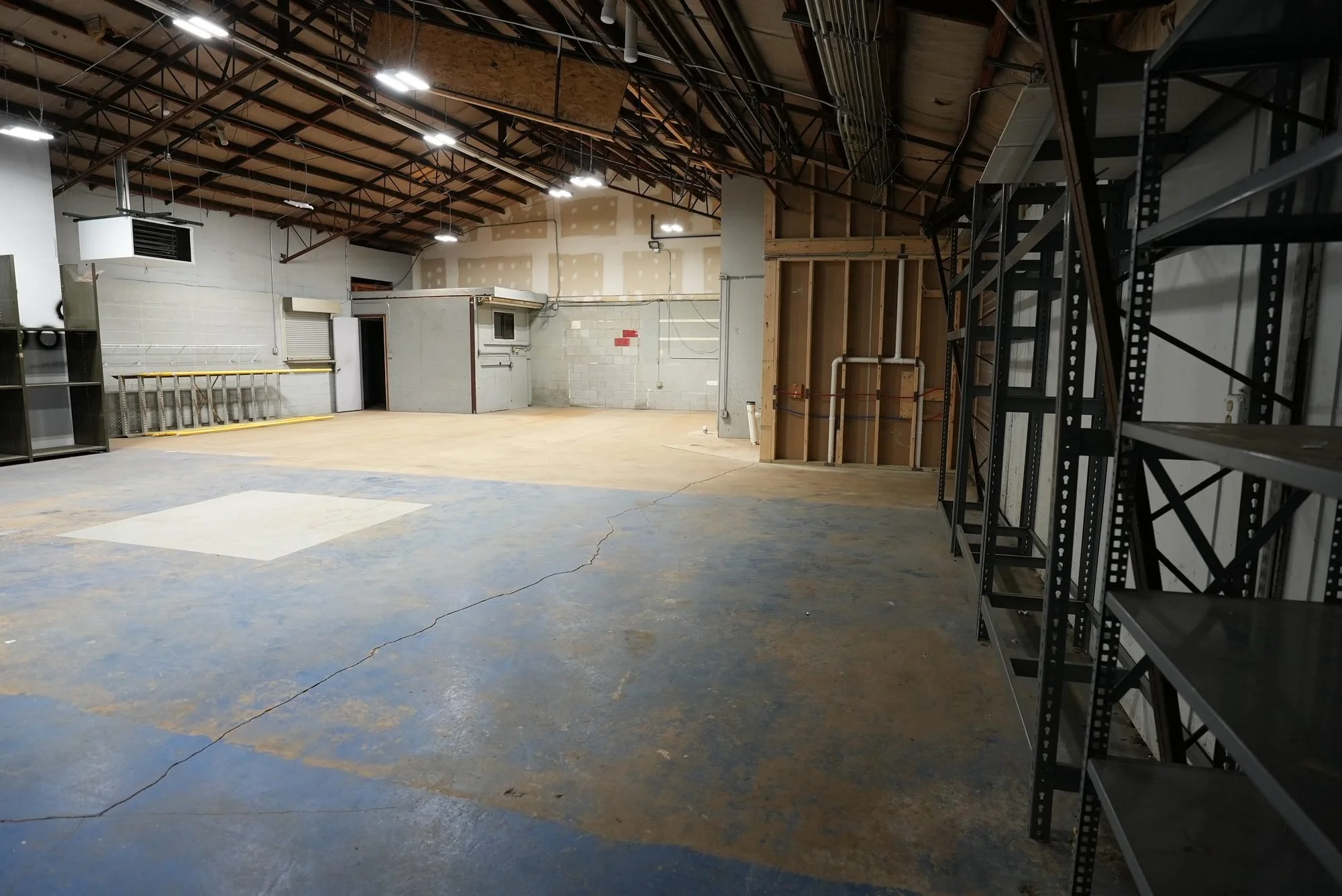 Empty indoor space with metal shelving on the right, unfinished walls, exposed ceiling with lighting, and a concrete floor with a large crack.