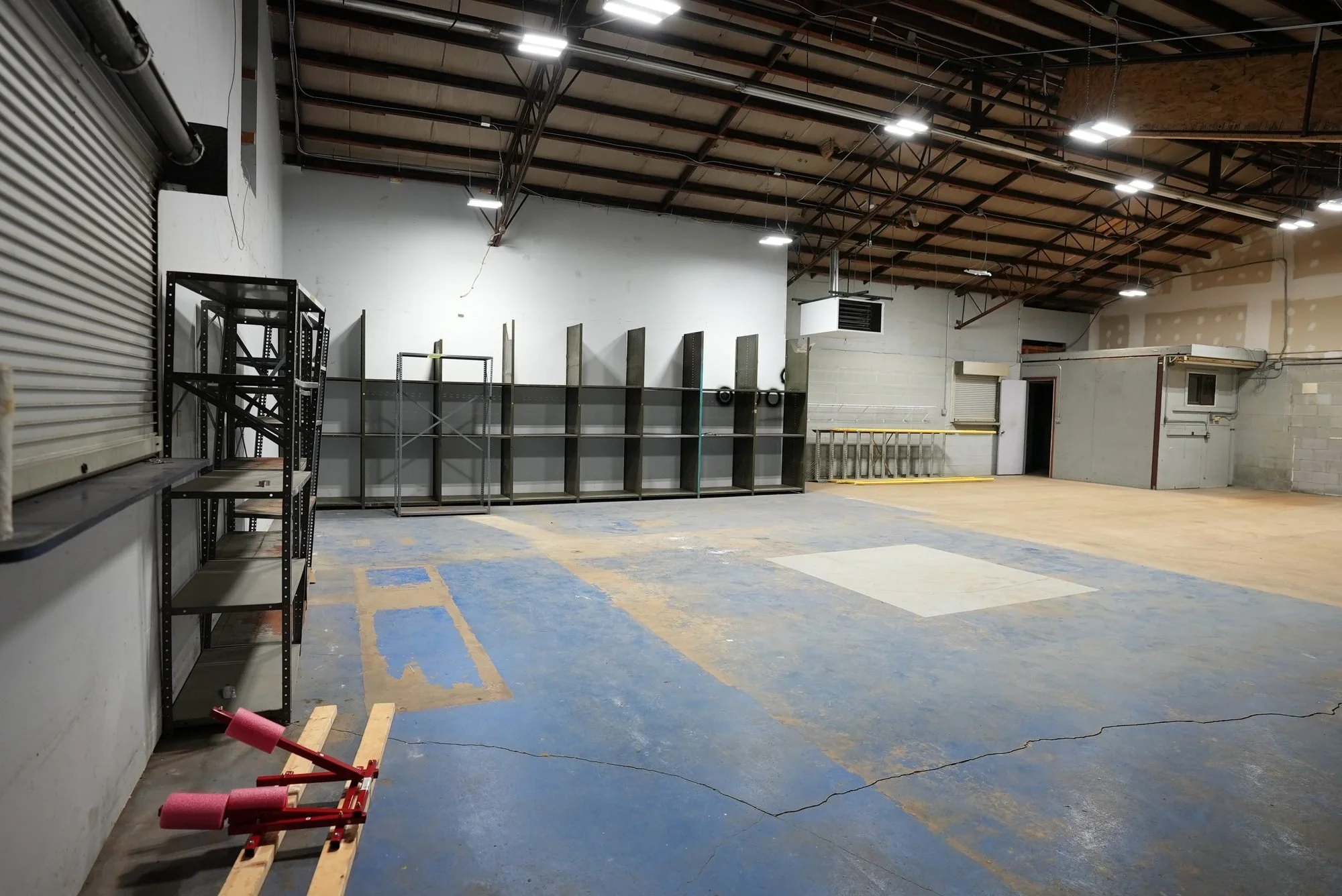Empty warehouse or industrial space with metal shelving, unfinished drywall, a rolling door, and a concrete floor with blue markings.