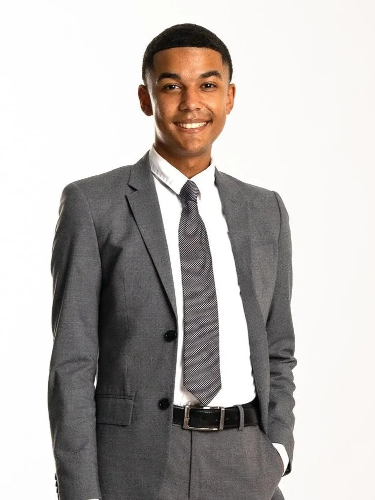 Isaiah Jobe-Winn wearing a gray suit with a white dress shirt and a dark tie, smiling, standing against a white background.