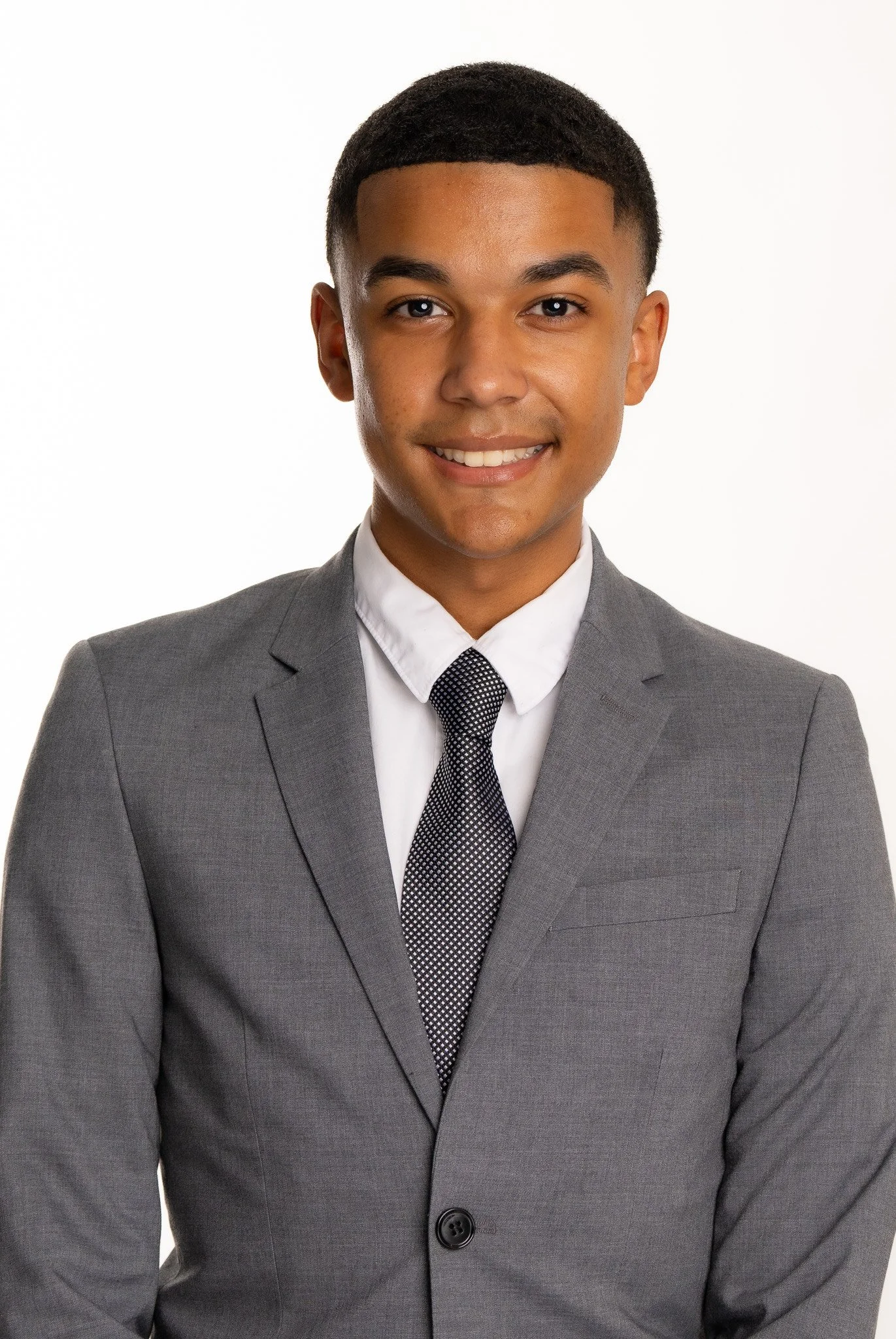 Isaiah Jobe-Winn wearing a grey suit, white shirt, and patterned black and white tie, smiling against a plain white background.