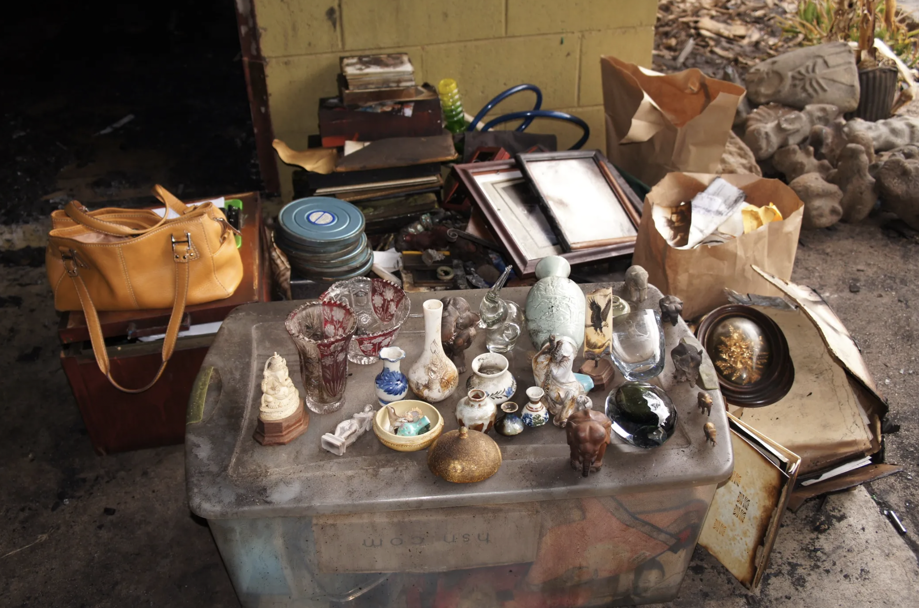 A table covered with vintage glassware, porcelain figurines, and small decorative objects, with a brown paper bag and picture frames behind it, in an outdoor setting with rocky ground and a brick wall.