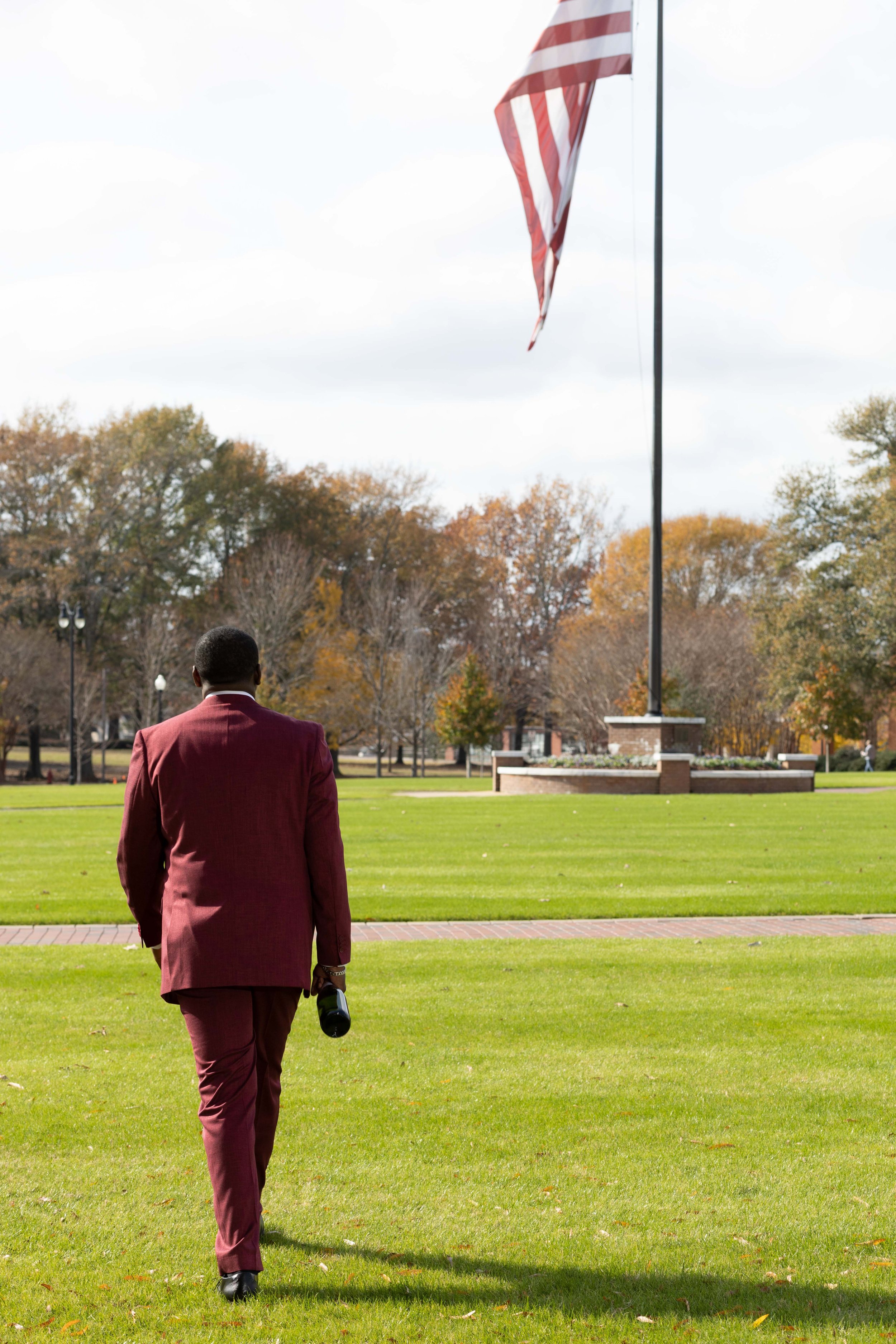 A man in a maroon suit walking toward a flagpole with an American flag in a park with autumn trees.