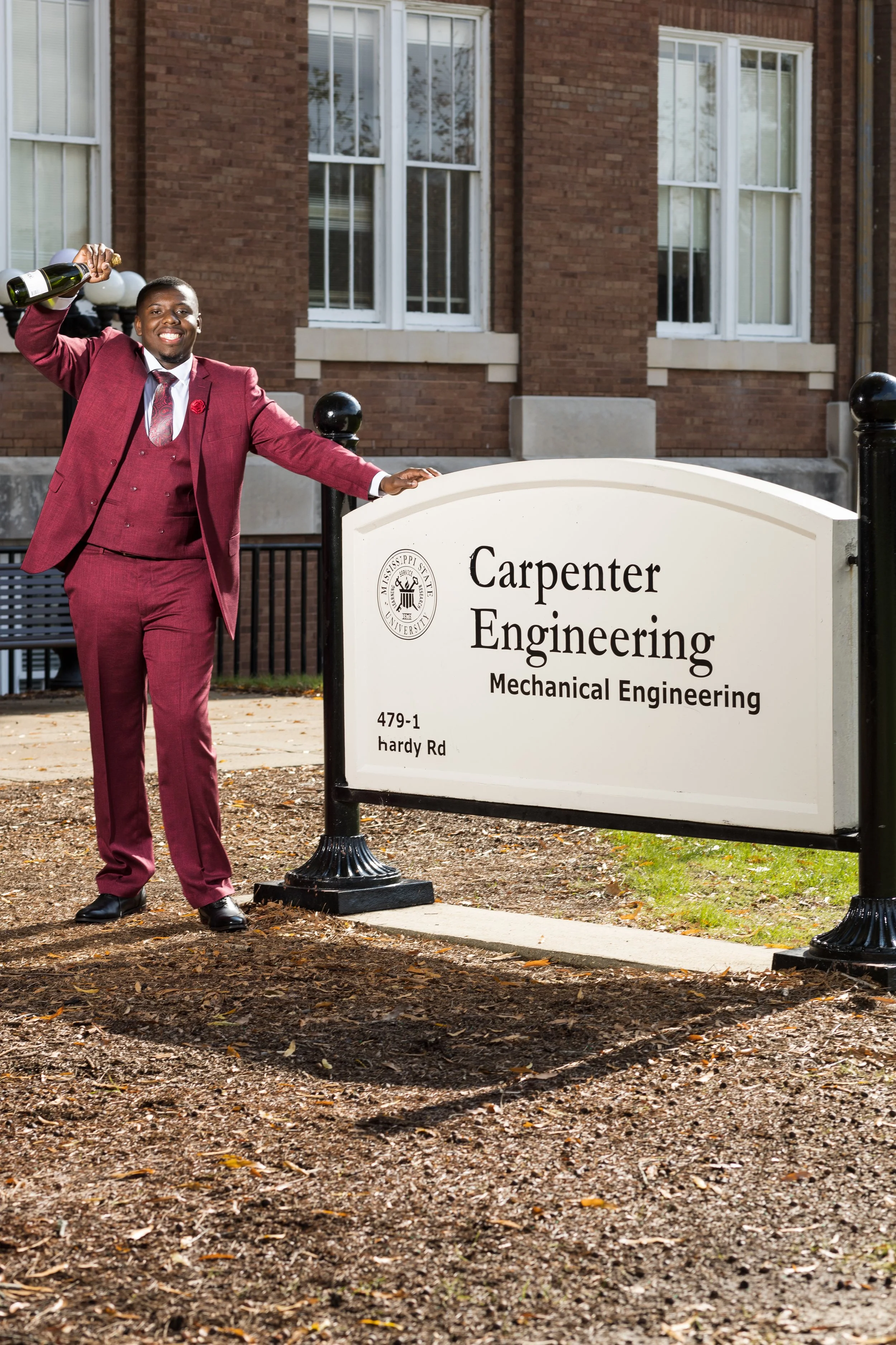 A smiling man in a red suit standing outside near a university sign for Mechanical Engineering, holding a bottle of champagne, in front of a brick building.