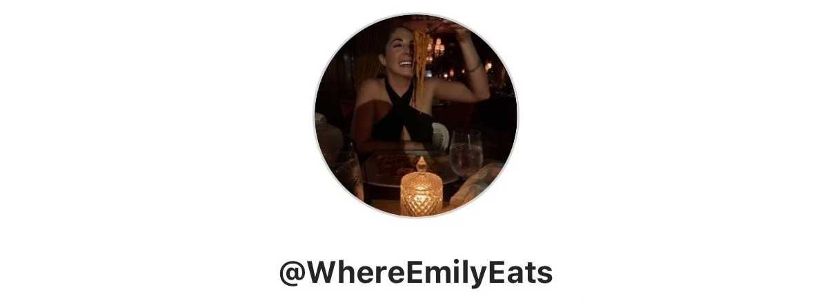A woman sitting at a restaurant table smiling, with a lit candle in front of her, in a dimly lit setting.