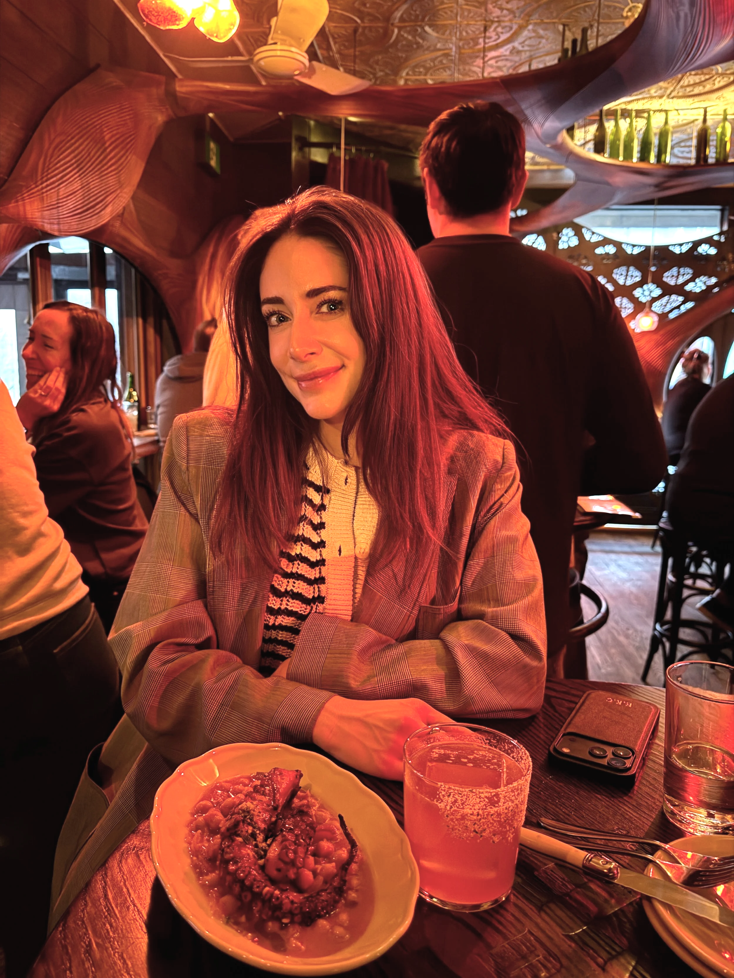 A woman with long brown hair and a checkered blazer smiling at a restaurant table with a plate of octopus tentacle in front of her, a glass of beer, and a glass of water, with other patrons and decorative interior in the background.