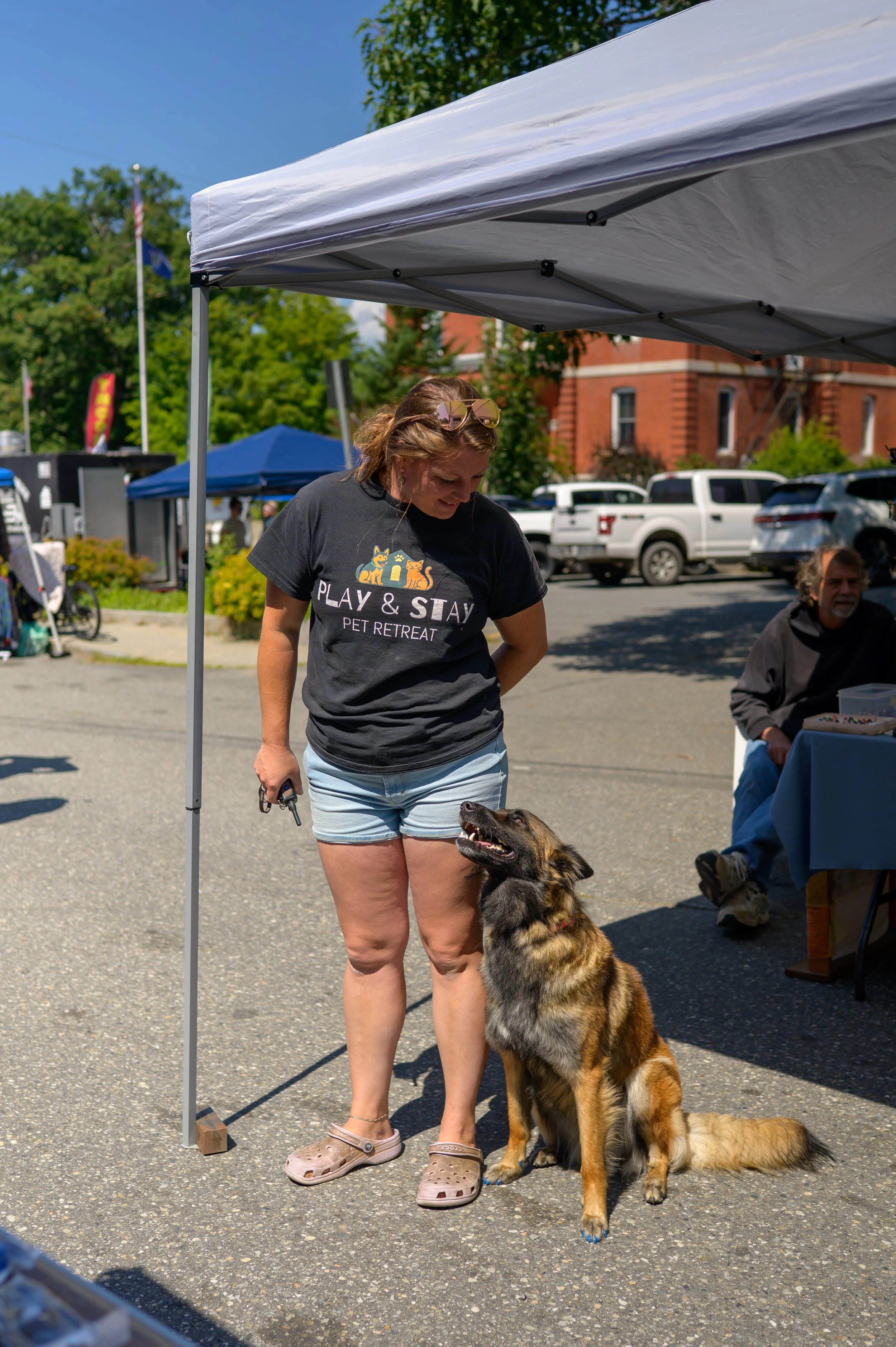 A woman with sunglasses on her head and wearing a dark t-shirt that says "PLAY & STAY PET RETREAT" is standing outdoors next to a large, fluffy German Shepherd dog. The woman is looking down at the dog, which is sitting and looking up at her. There are tents, parked cars, and a brick building in the background, suggesting a community or outdoor event on a sunny day.