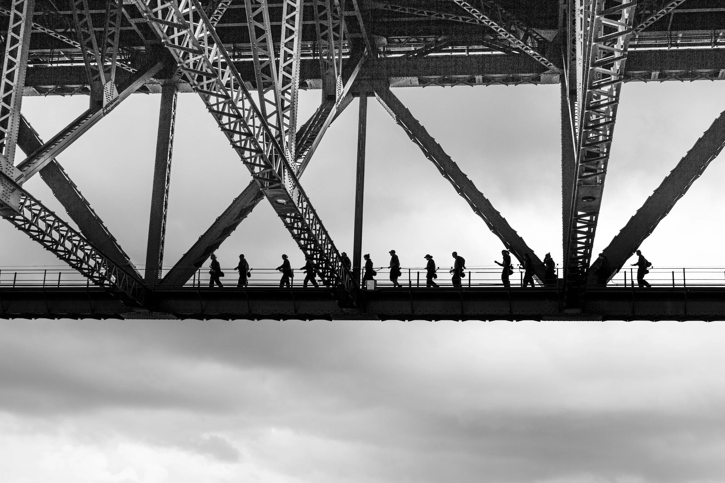 Black and white photo of people walking on a bridge beneath a large steel structure, likely a bridge or tower, with intricate metal framework.