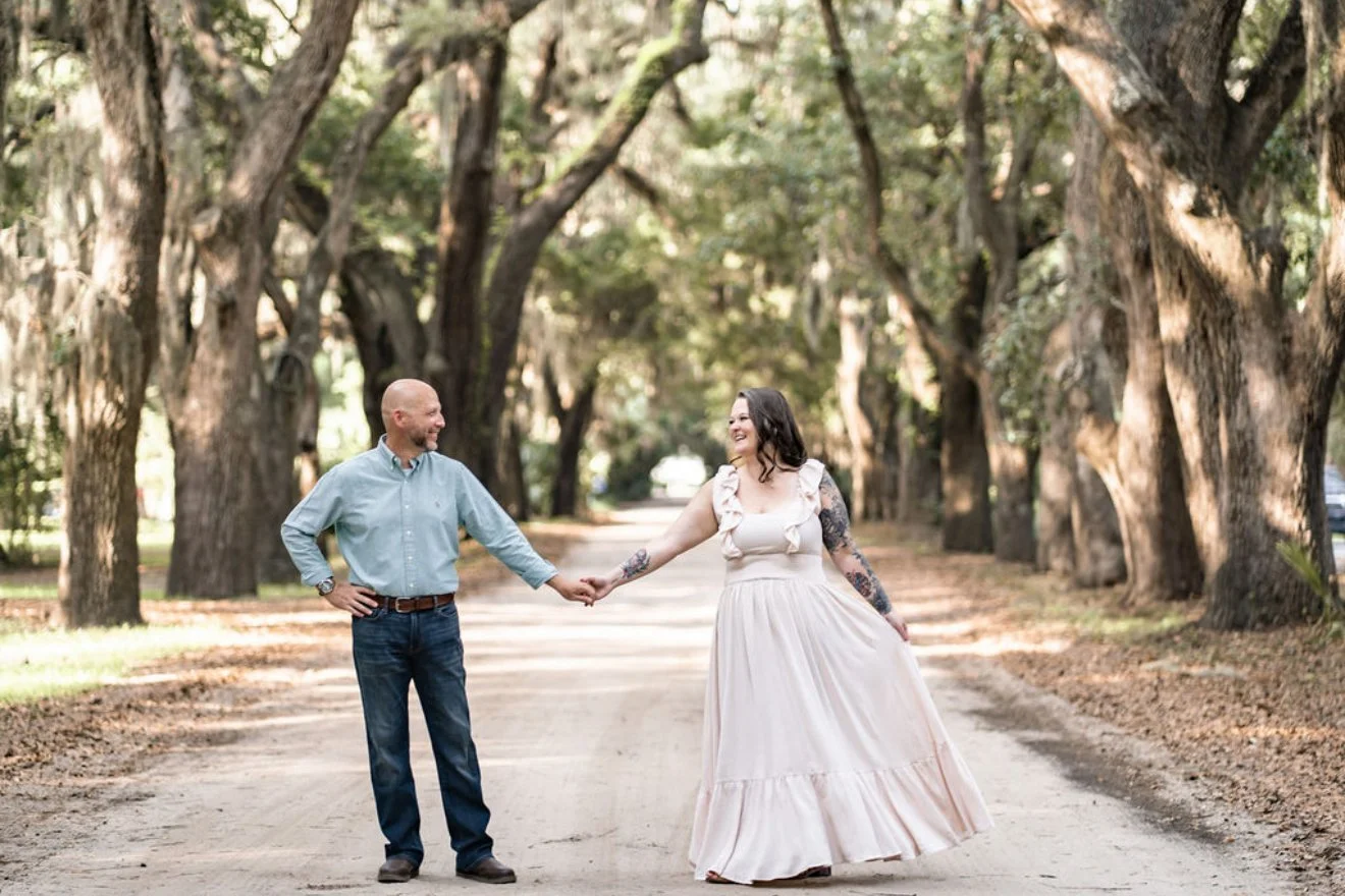 A couple holding hands and smiling at each other in a tree-lined outdoor setting.