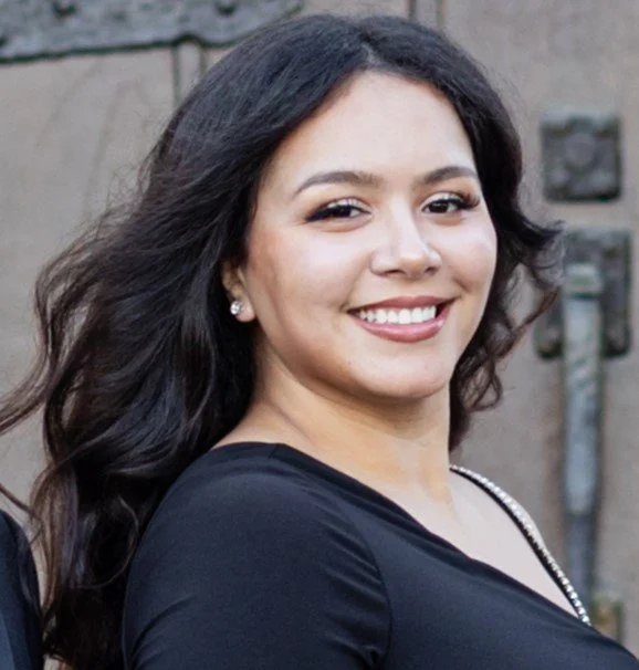 A woman with long dark hair smiling outdoors, wearing a black top, earrings, and a necklace.
