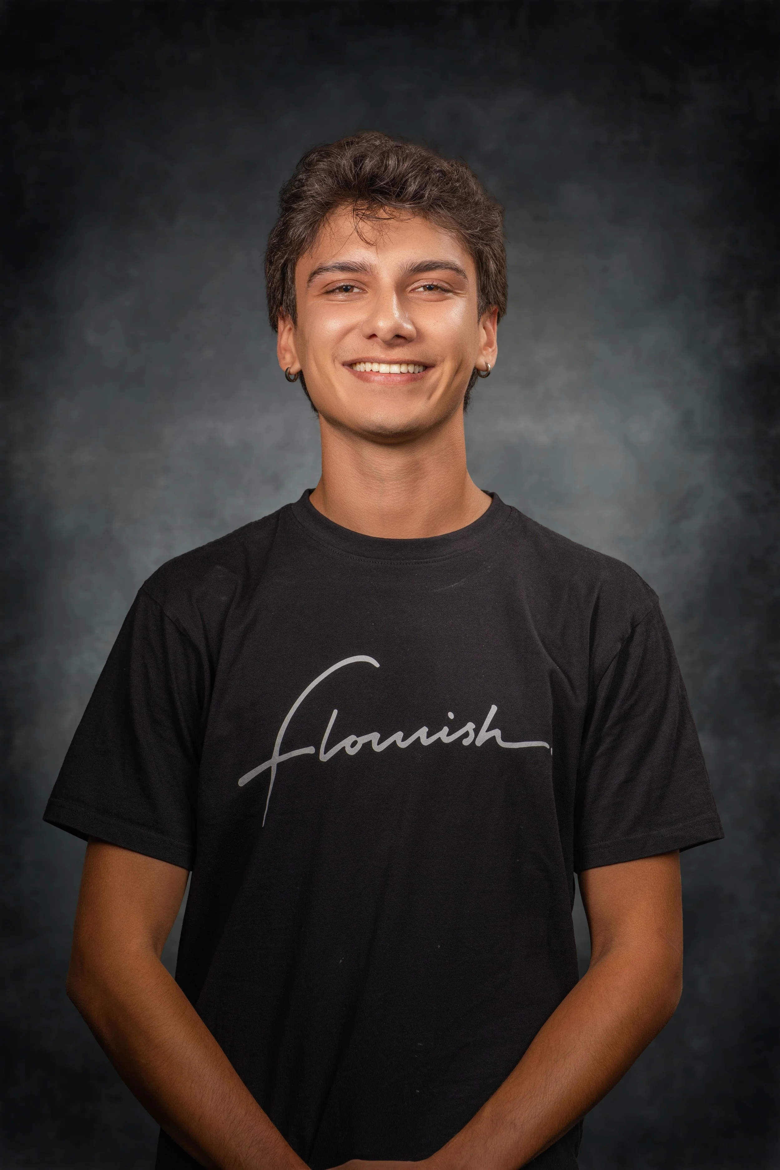 A young man with short brown hair, earrings, and a slight smile, wearing a black T-shirt with the word 'flourish' written on it, is standing against a dark, mottled background.
