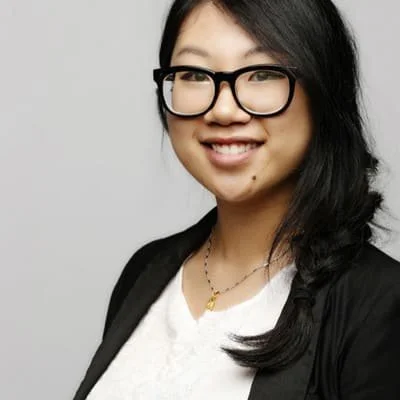 A young woman with black hair, wearing glasses, a white top, and a black blazer, smiling at the camera.