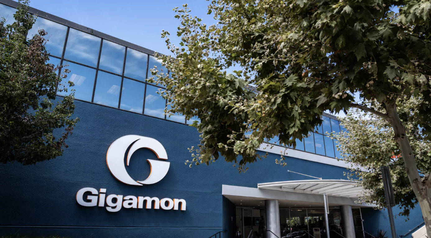 Exterior view of Gigamon office building with a large blue sign displaying the Gigamon logo and name, surrounded by trees and a blue sky.