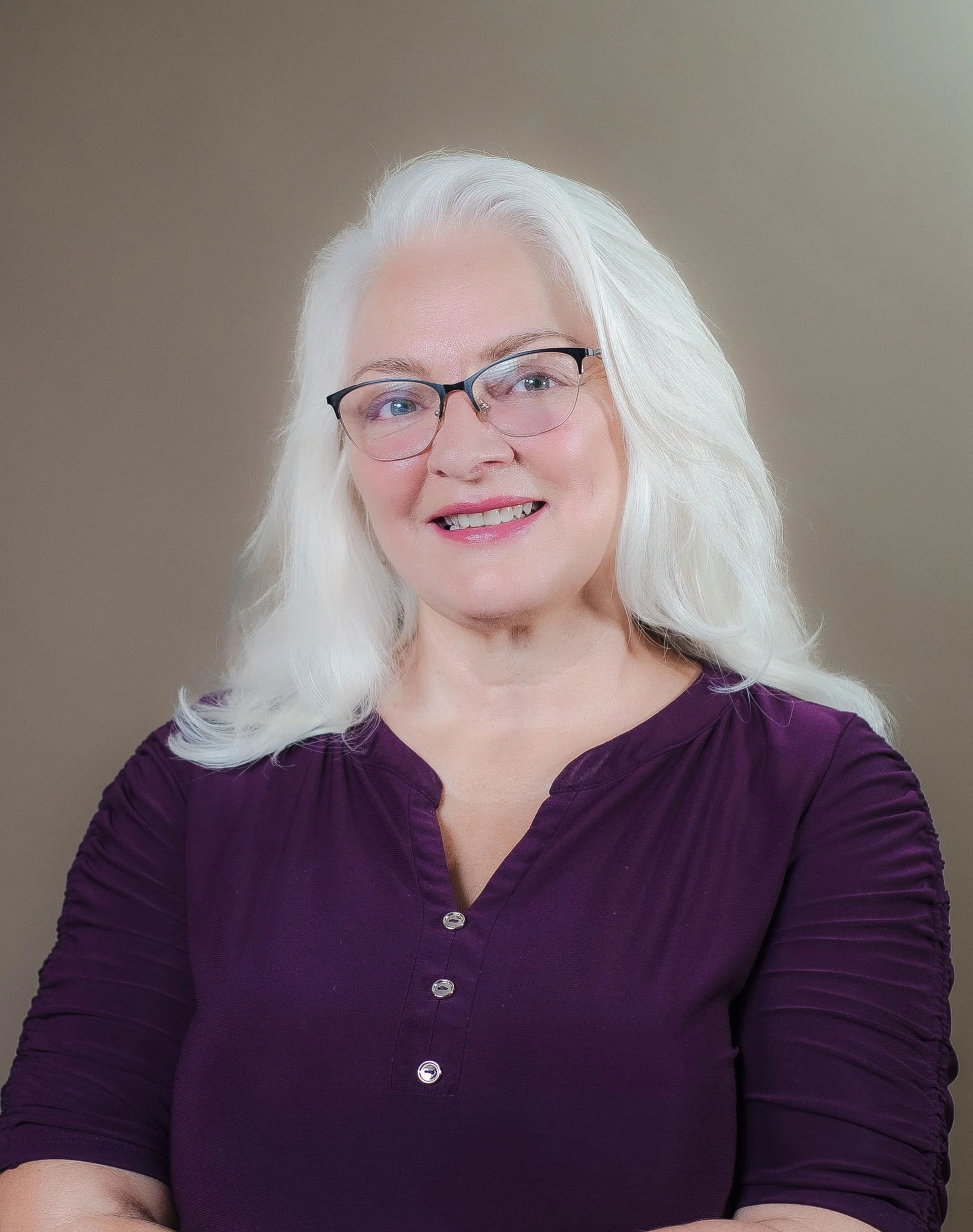 Close-up of a smiling woman with white hair and glasses wearing a purple top.