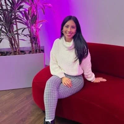 A woman with long dark hair smiling, wearing a white sweater and gray checkered pants, sitting on a red sofa in front of purple lighting and potted plants.