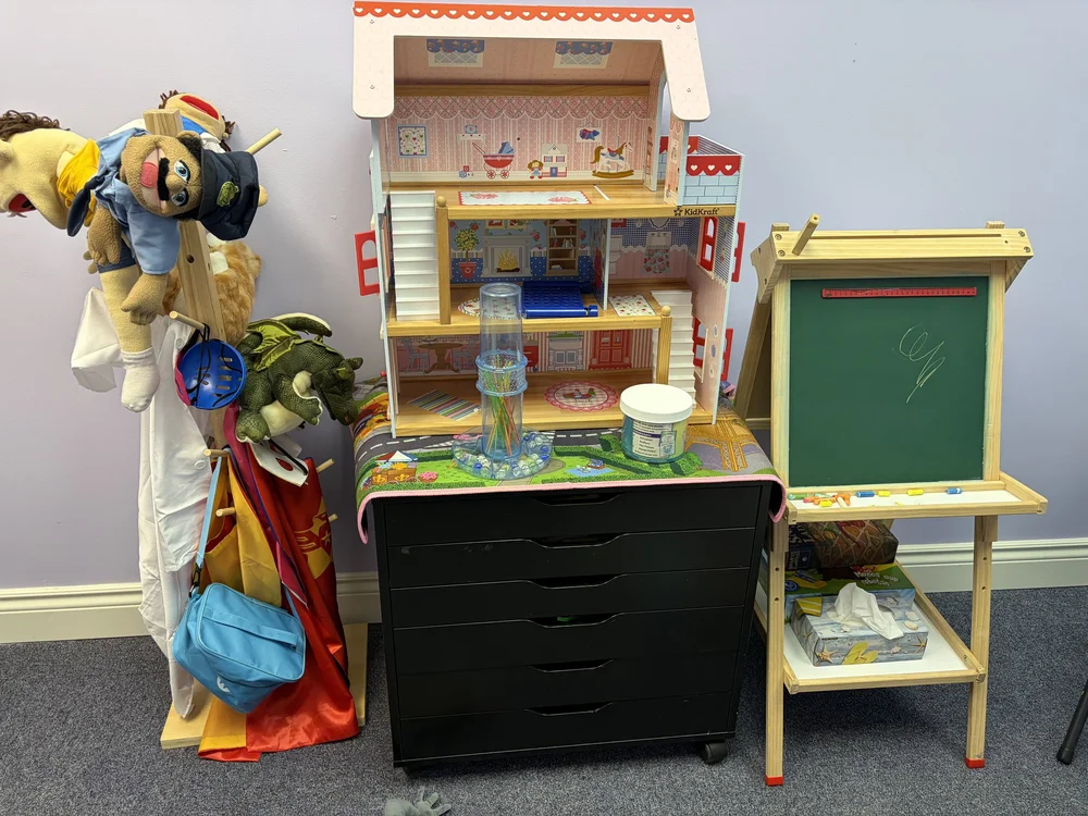 A dollhouse, puppets, dress-up clothes and a small easel chalkboard in Riverway's play room