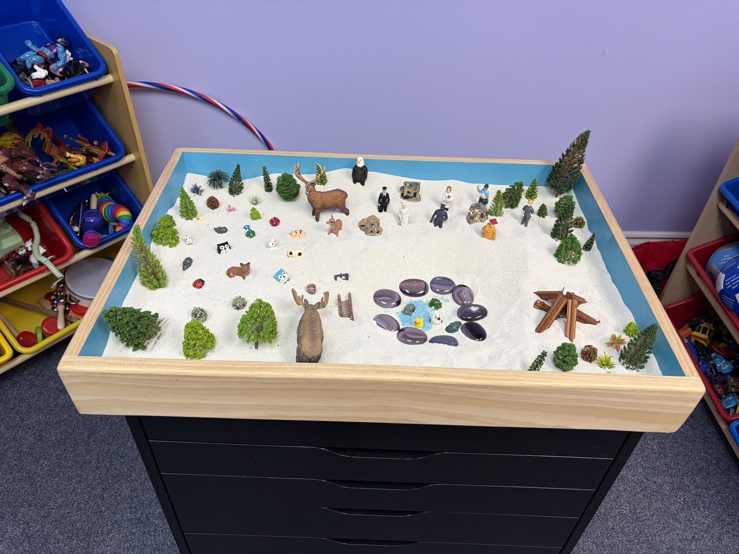 A sand tray in Riverway's play room, set up with small animal, tree and people figures on white sand
