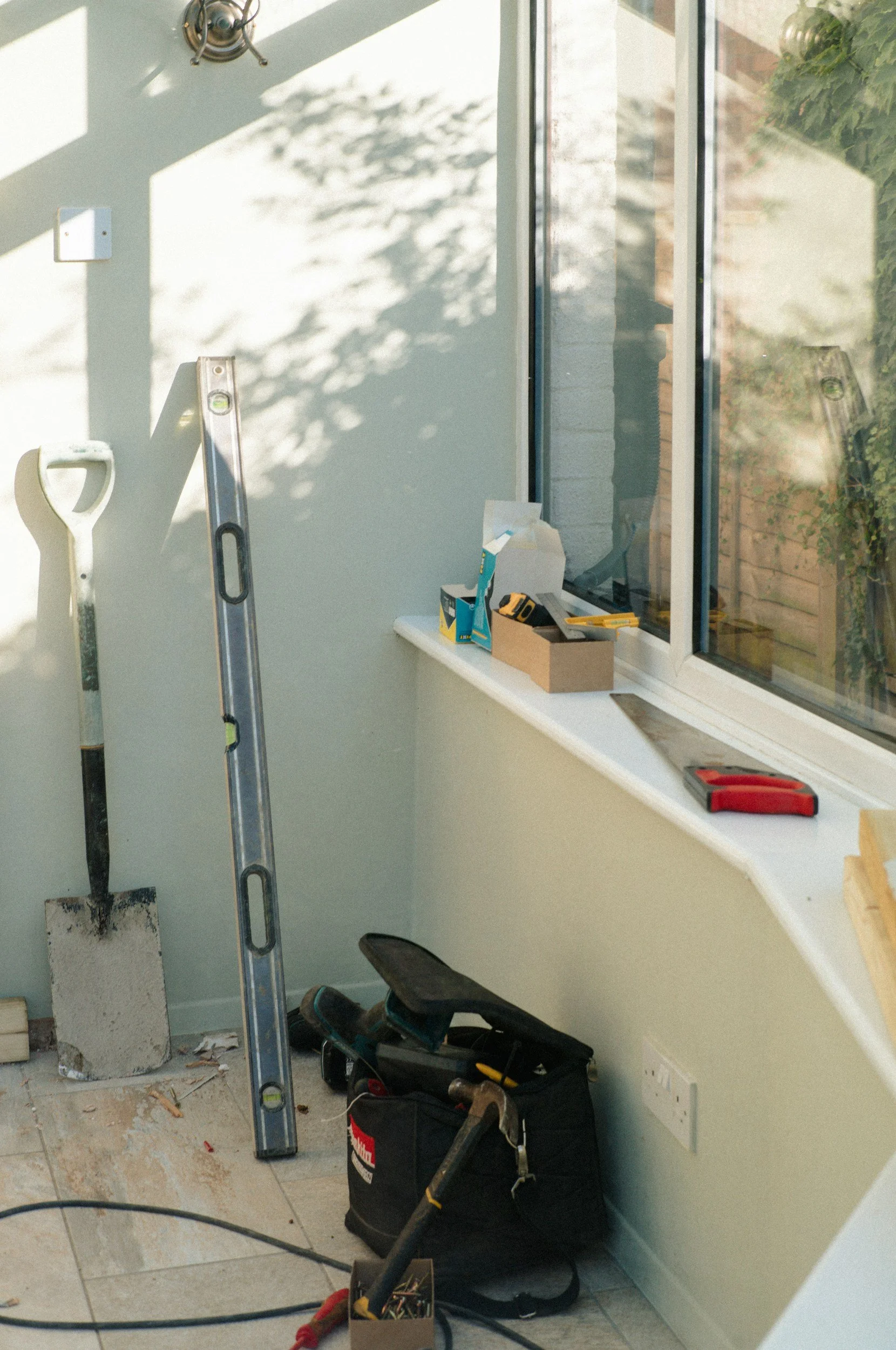 A partially completed interior wall with tools and equipment, including a level, a shovel, a toolbox, and a bag, near a window and electrical outlet.