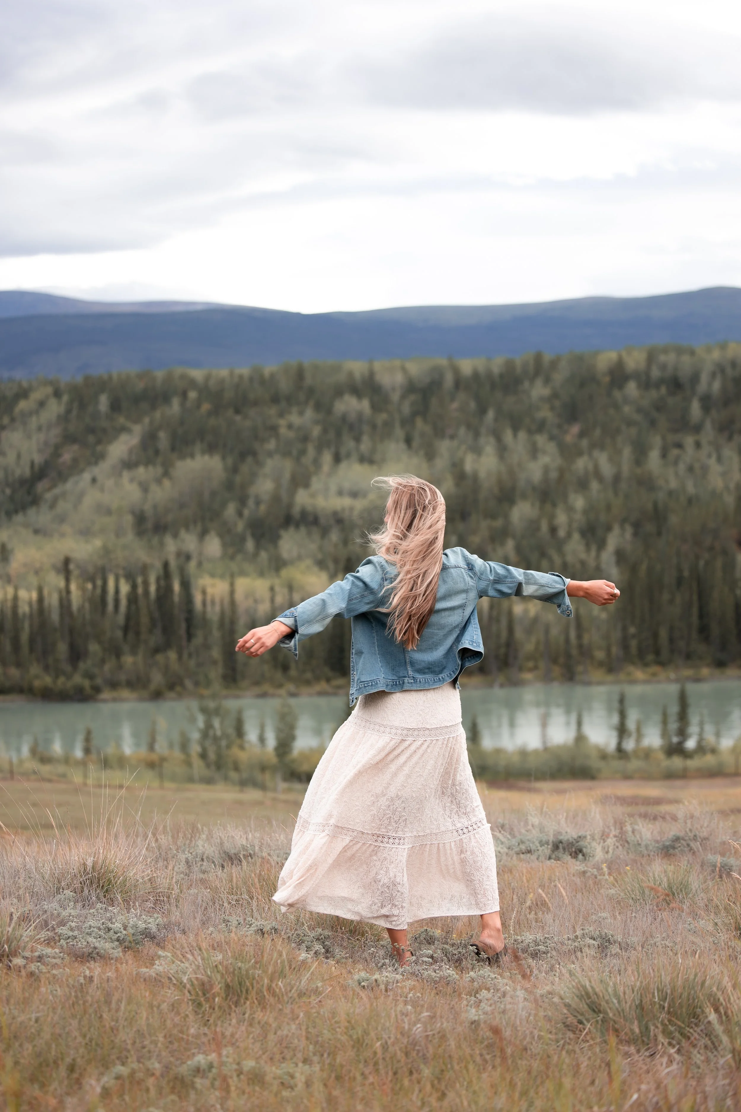 A woman with long blonde hair standing in a field near a body of water with mountains in the background, her arms outstretched in a carefree pose.