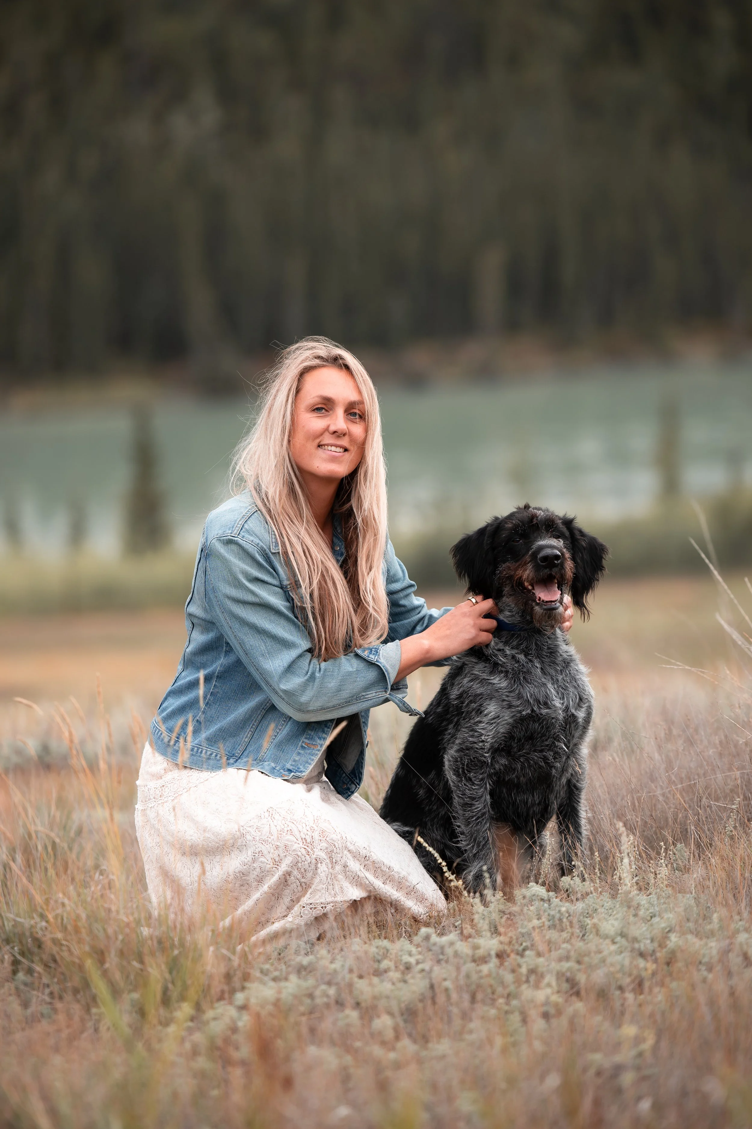 A woman with long blonde hair wearing a denim jacket and a white lace dress kneeling in a field with a black and gray dog, both looking at the camera near a lake or river with a forested background.
