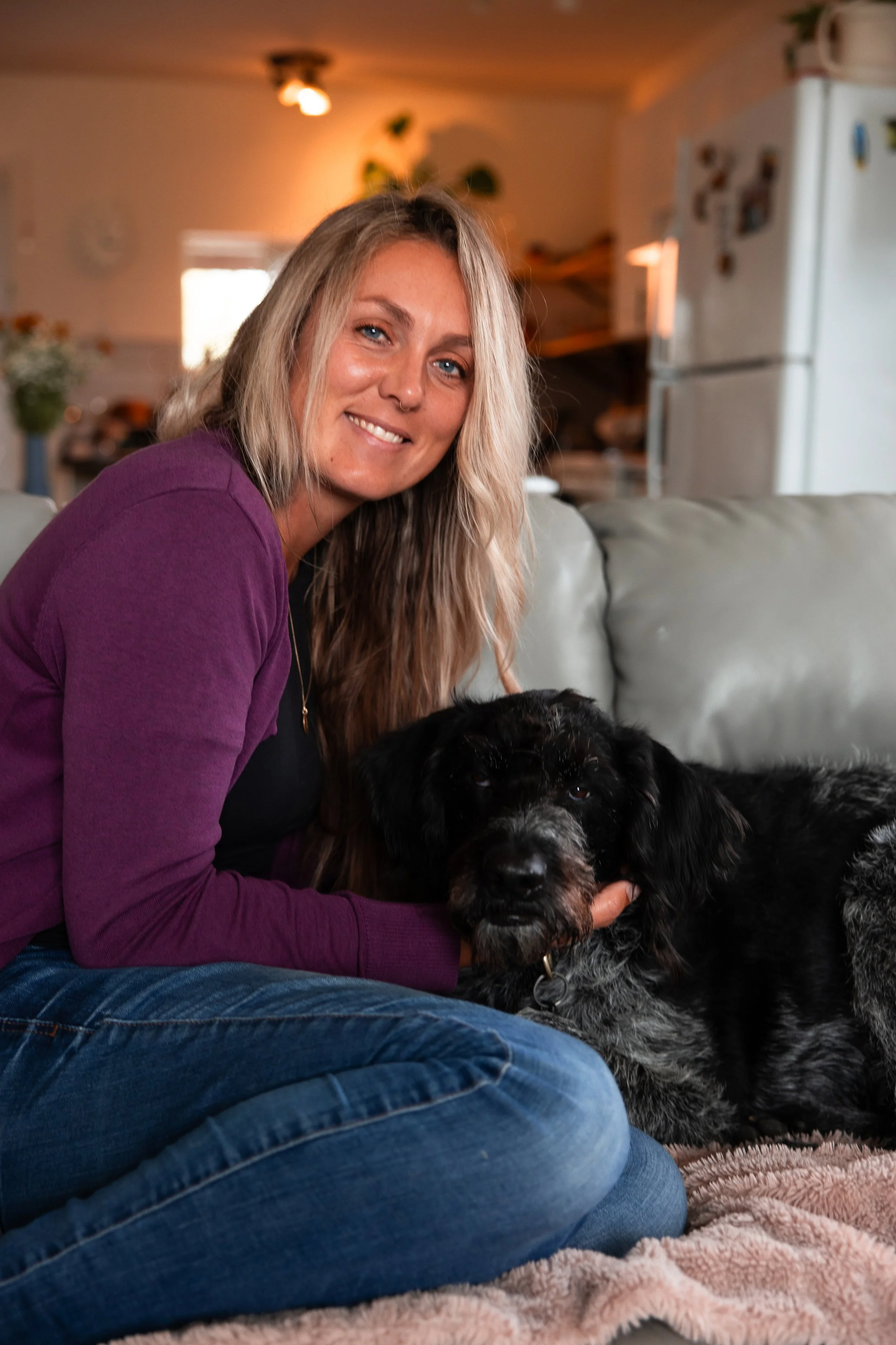 A woman with long blonde hair and a purple sweater sitting on a couch with a black and white dog, in a cozy living room.