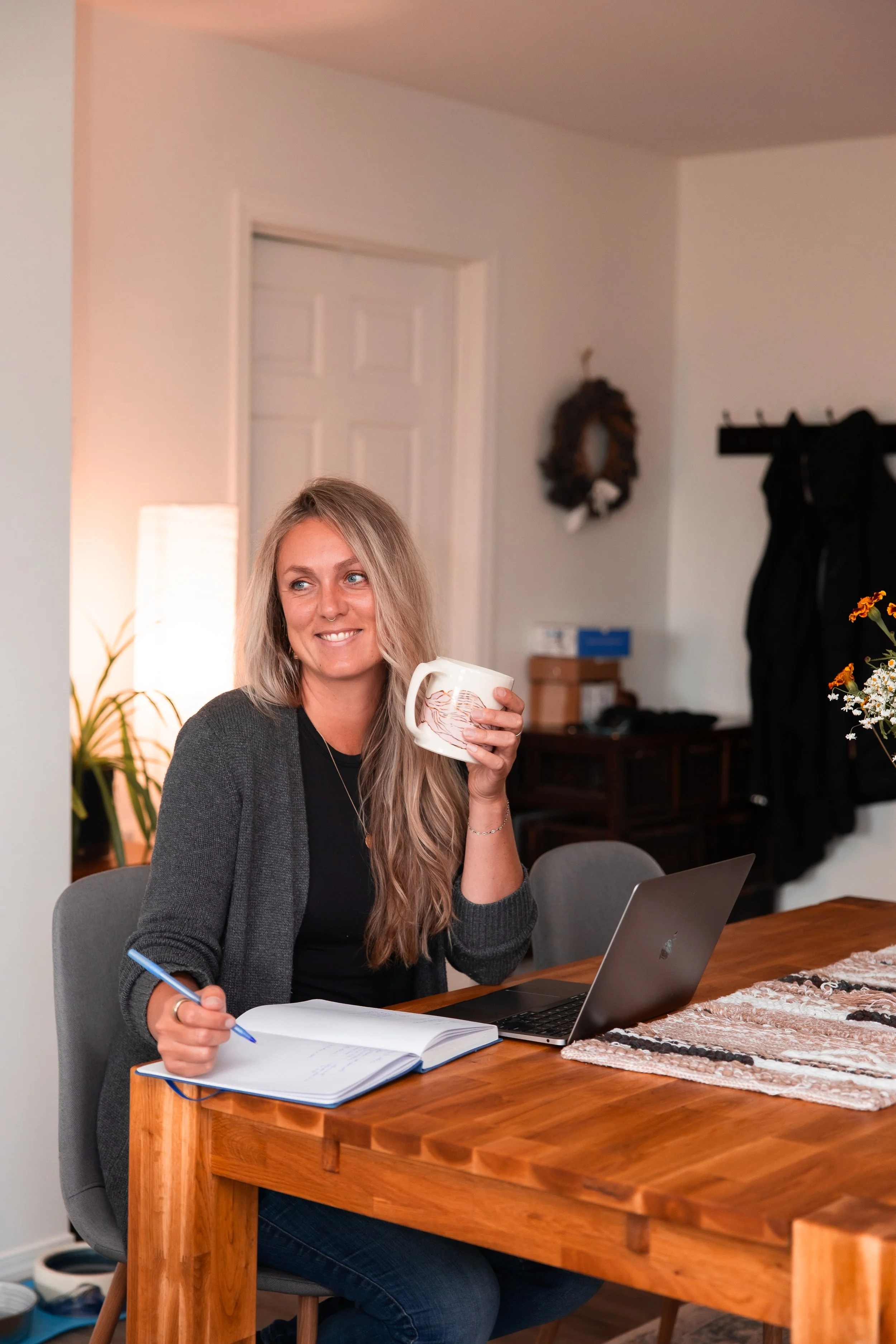 A woman with long blonde hair, wearing a black shirt and grey cardigan, is sitting at a wooden table, smiling, holding a mug in her right hand, and writing in a notebook with a blue pen in her left hand. There is a laptop open on the table in front of her, and she is sitting in a grey chair. The background shows a white wall with a decorative wreath, a coat rack, and some furniture.