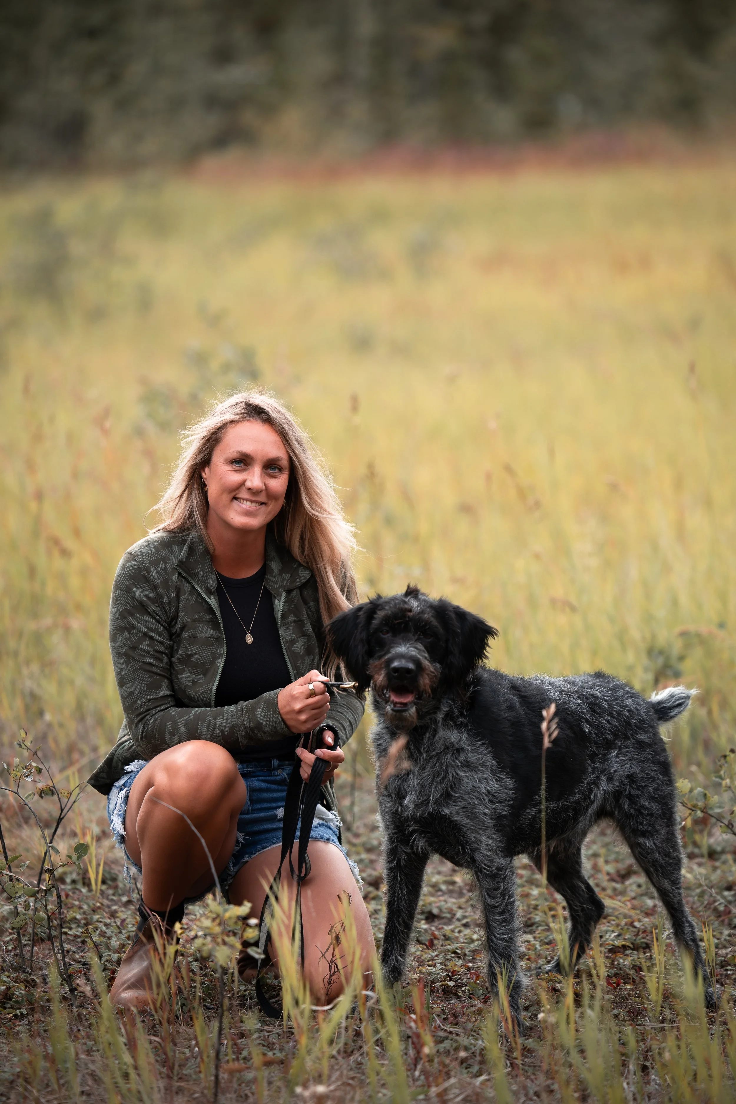 A woman with long blonde hair kneeling in a grassy field, holding the leash of a large black and gray Australian Shepherd dog with a fluffy coat and happy expression.