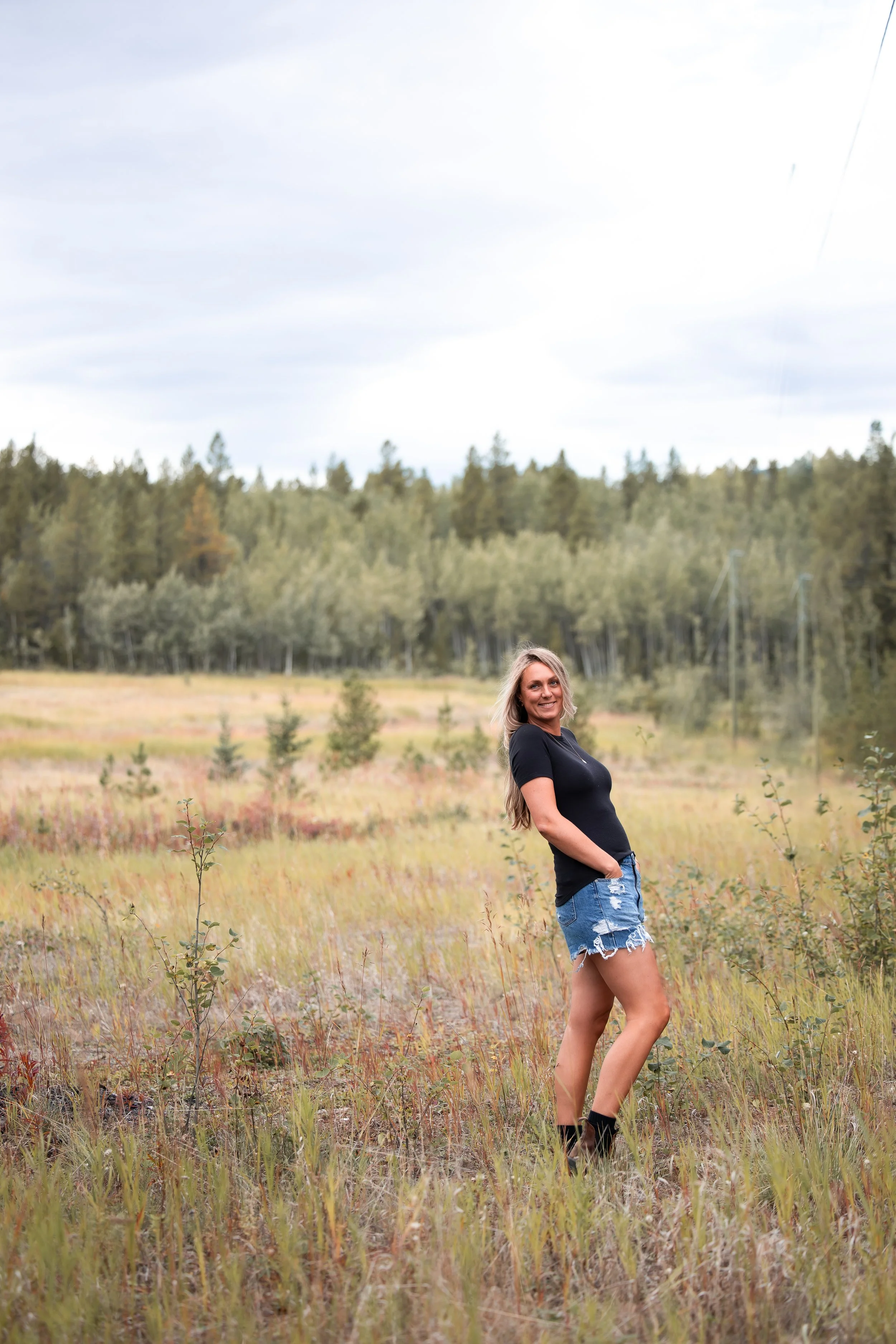 A woman with long blonde hair, wearing a black t-shirt and ripped denim shorts, standing in a field of wild grass with trees in the background under a cloudy sky.