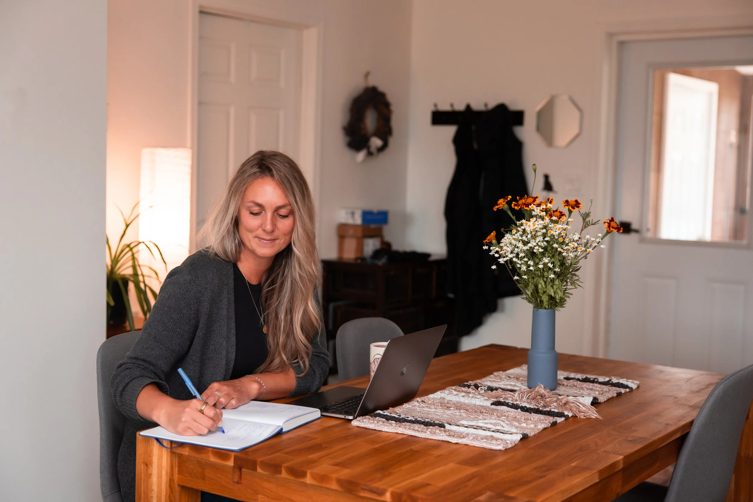 A woman with long blonde hair sitting at a wooden table, writing in a notebook with a pen. There is a laptop, a mug, and a vase with yellow, white, and orange flowers on the table. The background shows a white wall, a door, a mirror, a coat rack, and a wreath hanging on the wall.