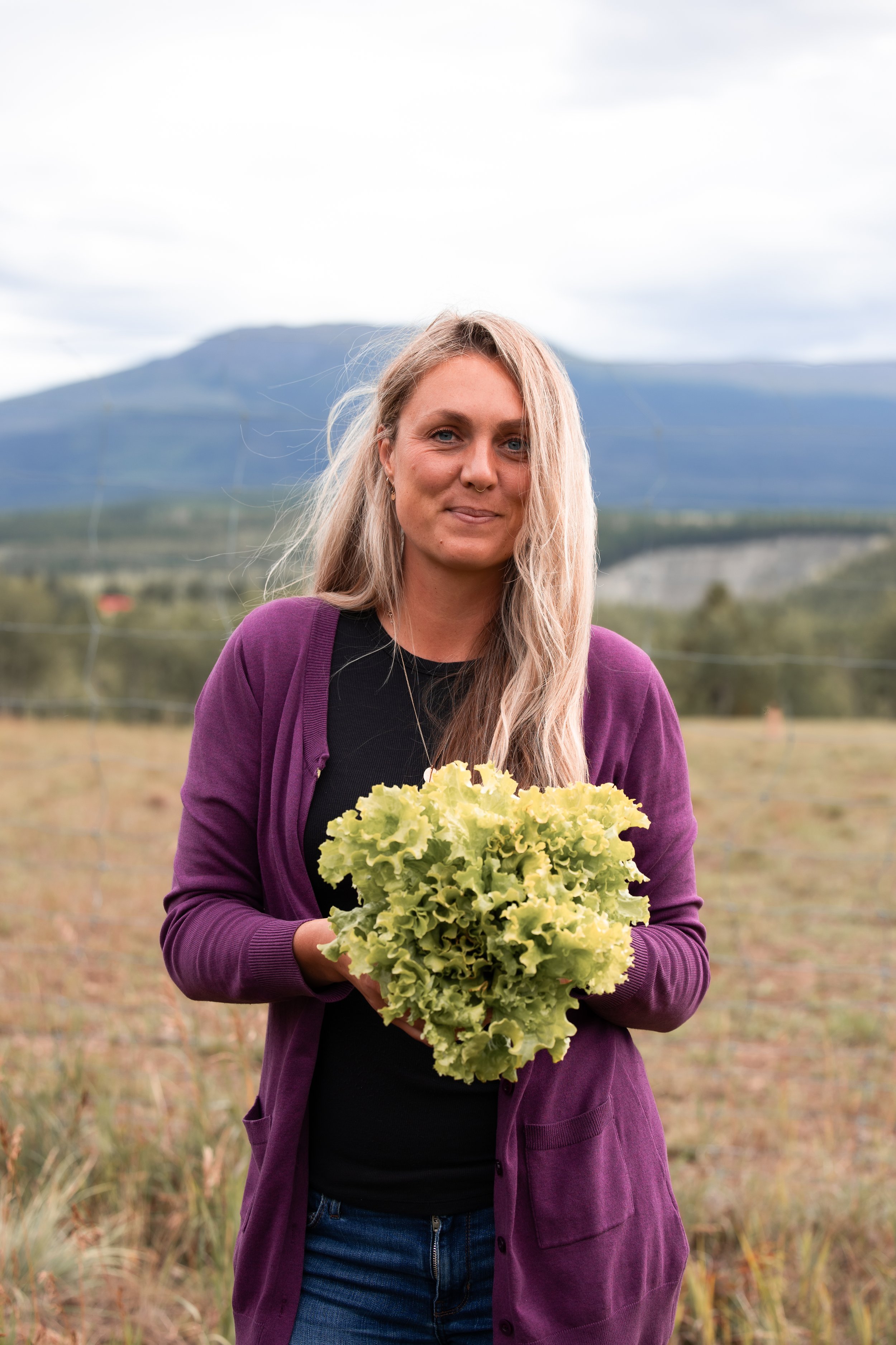 A woman with long blonde hair, wearing a black shirt and purple cardigan, holding a bunch of leafy green lettuce, standing outdoors in a field with mountains and cloudy sky in the background.