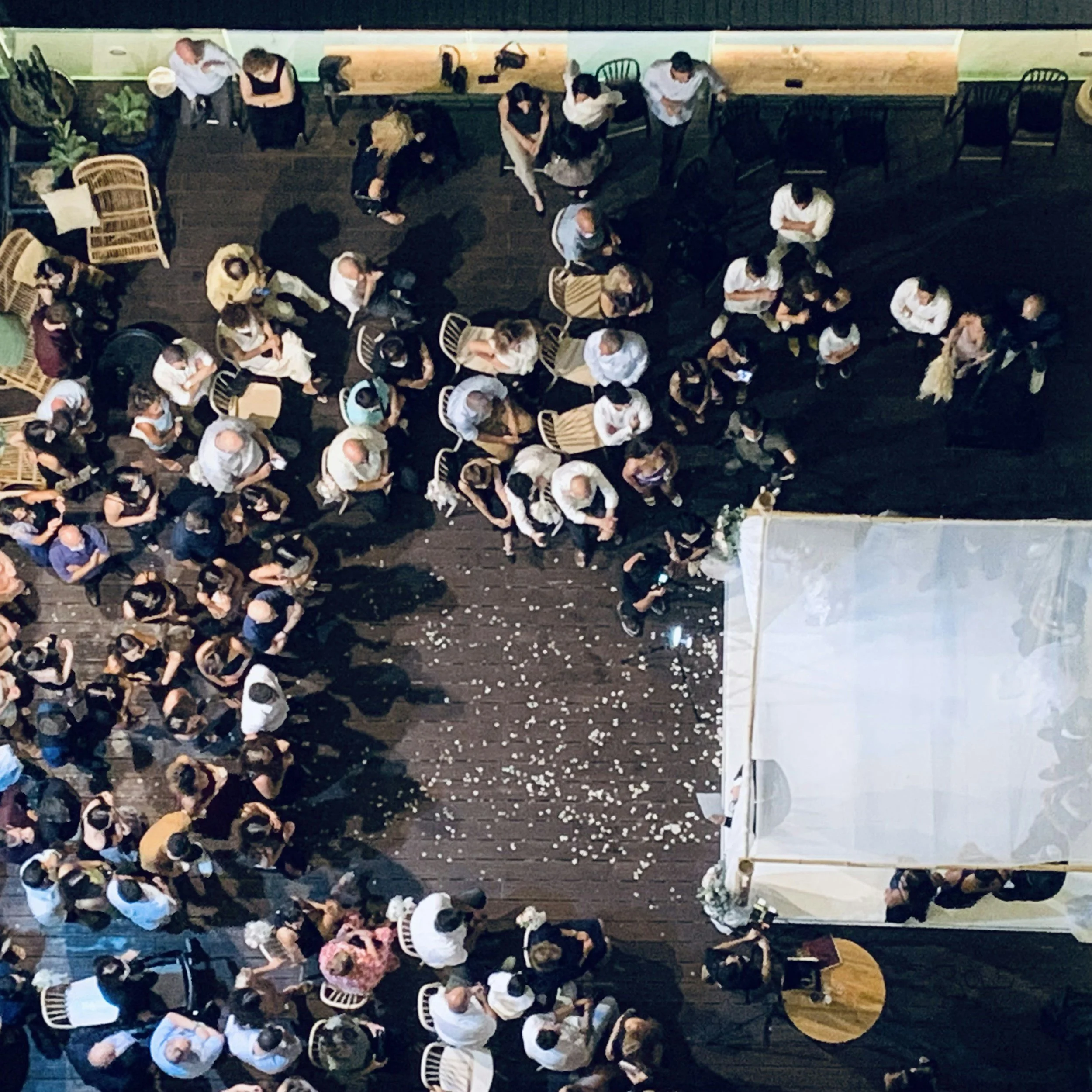 An overhead view of a large group of people sitting and standing around tables at a social event or restaurant, with some people talking and some using their phones.