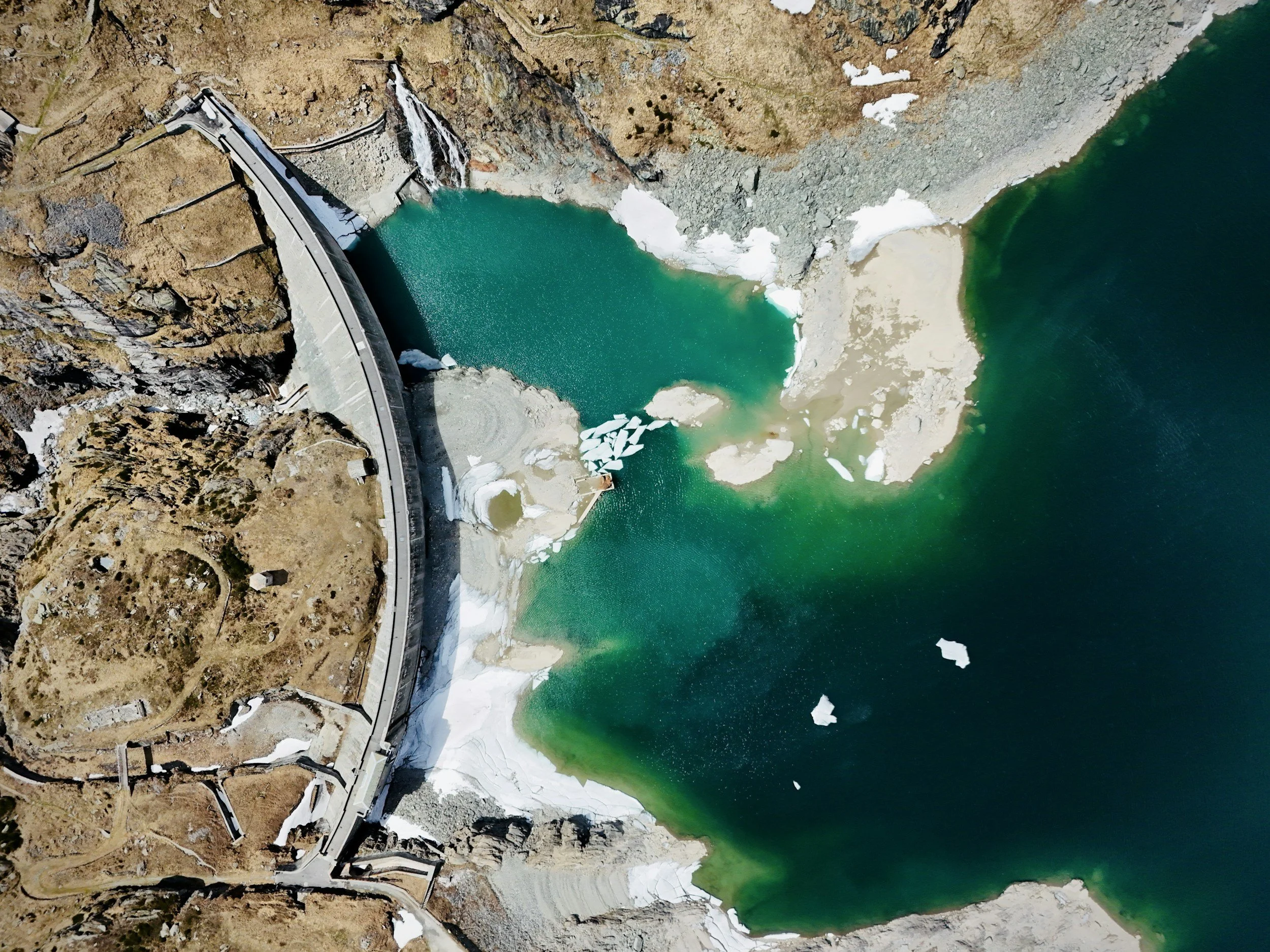 Aerial view of a dam reservoir with a curved shoreline, surrounded by brown and rocky terrain, with patches of snow and ice on the edges of the water.