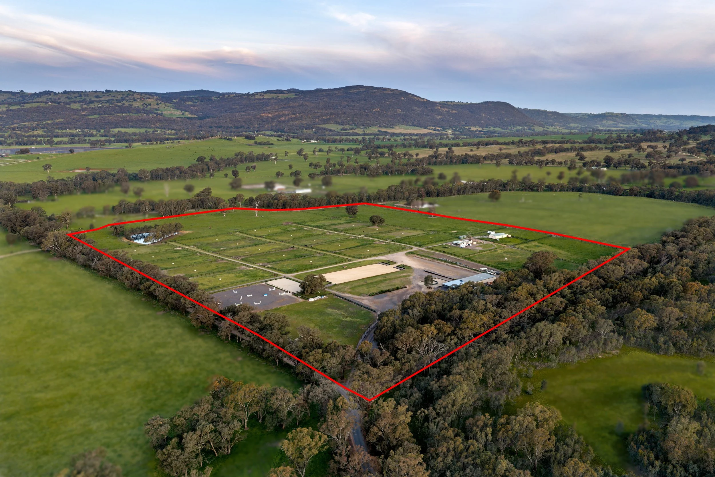 Aerial view of a large fenced property with green fields, trees, buildings, and a pond, set in a rural landscape with mountains in the background.